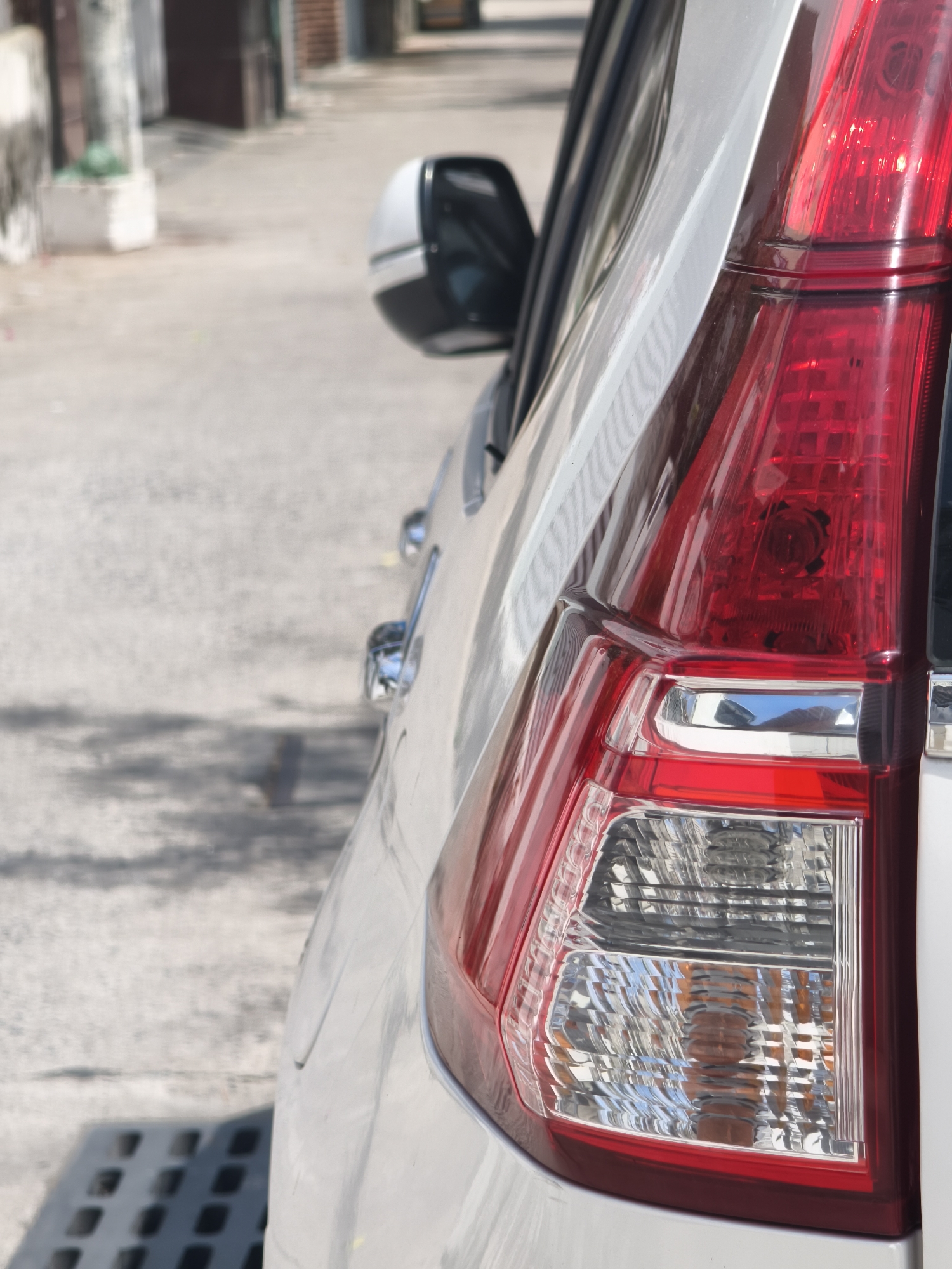 A close-up, side-view, outdoor shot focuses on the rear passenger-side quarter panel and taillight assembly of a white SUV parked on a textured, light-colored asphalt surface, possibly a street or parking lot. The taillight is a prominent red and clear lens with visible internal reflectors and bulbs. A chrome accent piece is visible below the red portion of the taillight. The body of the SUV is white and shows a smooth, curved surface. In the background, a narrow, muted-colored street or alleyway is visible, with the blurry outlines of buildings and a dark green object, possibly foliage in a planter, on the left side. The image is characterized by soft, natural lighting casting shadows on the ground. The overall scene is devoid of people or any explicit action. The context of Nha Trang, Vietnam, is not visually identifiable within the image itself.