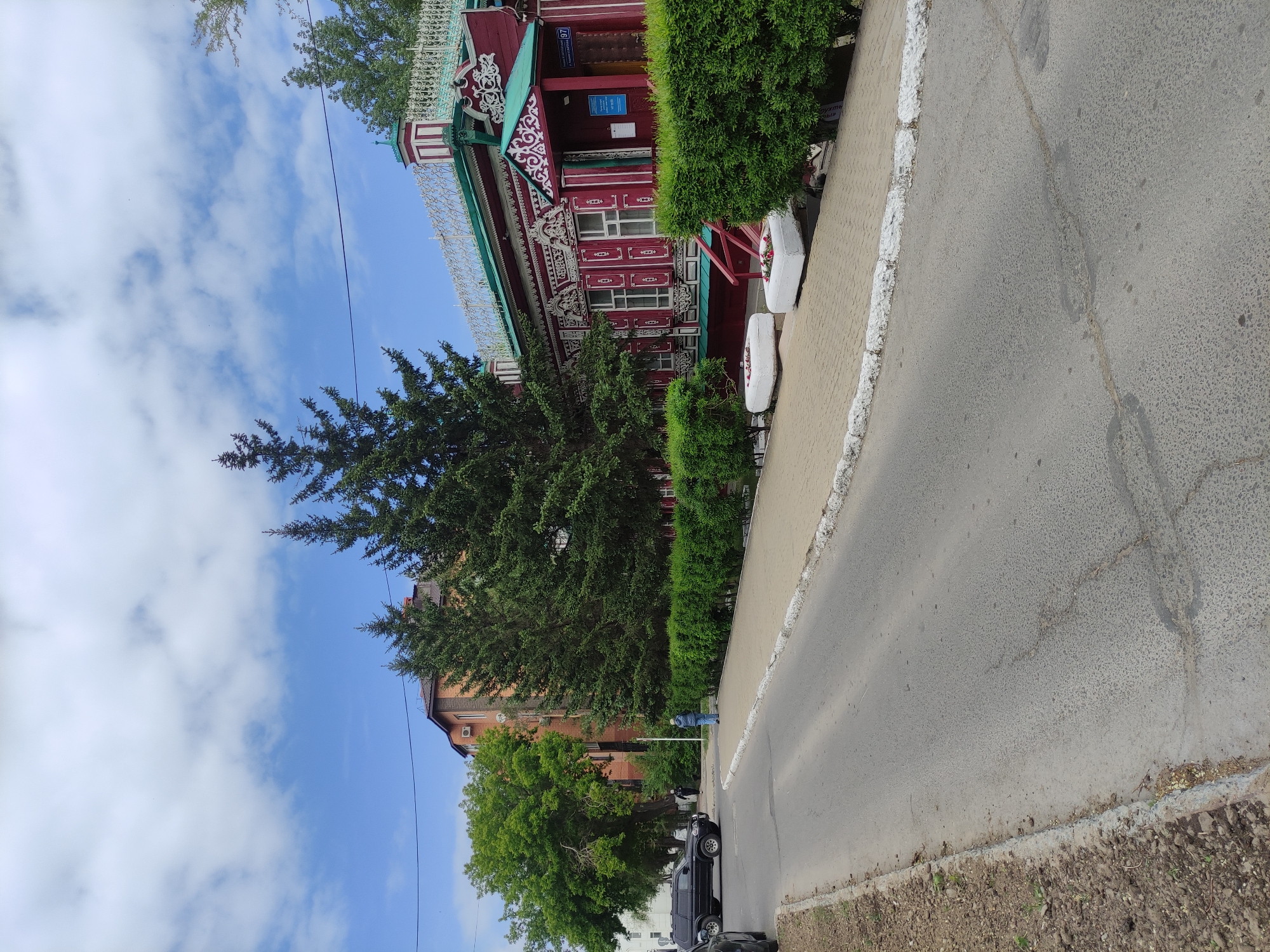 The image shows a street scene in Pavlodar, Kazakhstan. On the left, a traditional, ornately decorated wooden house, painted in red, green, and white, stands next to a tall evergreen tree. A trimmed hedge separates the house from the sidewalk. To the right, the asphalt street extends into the distance. A black SUV is parked further down the street, near a person standing on the sidewalk. The sky is visible above, with scattered clouds and blue patches. A few overhead power lines cut through the sky. The image is slightly tilted.