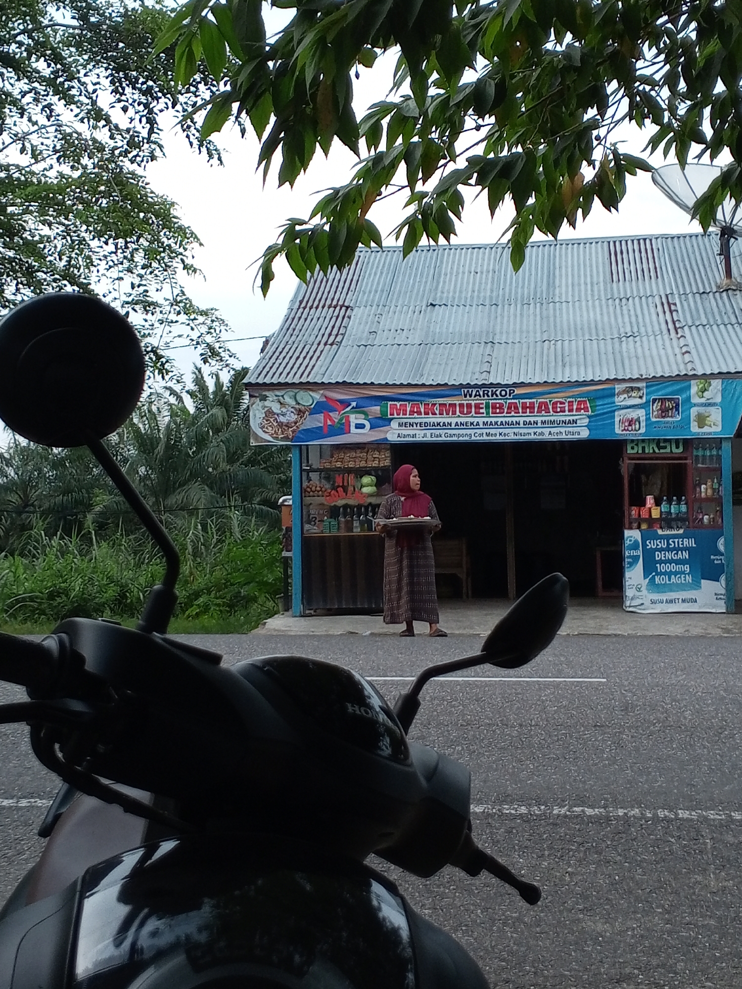 A low-angle, outdoor shot captures a roadside food stall named "WARKOP MAKMUE BAHAGIA." The stall, with a corrugated metal roof, is adorned with a banner showcasing a noodle dish and the text "MENYEDIAKAN ANEKA MAKANAN DAN MINUMAN," along with an address. To the left of the stall, a woman wearing a maroon hijab and a long, patterned dress is seen holding a tray with food. In front of the stall are various items displayed for sale, including bottled drinks. To the right of the stall, a poster advertises "SUSU STERIL DENGAN 1000mg KOLAGEN." The foreground of the image is dominated by the silhouette of a motorcycle, with its handlebars and mirror visible. The background consists of lush green vegetation, including palm trees, and a cloudy sky. The scene appears to be located on a paved road.
