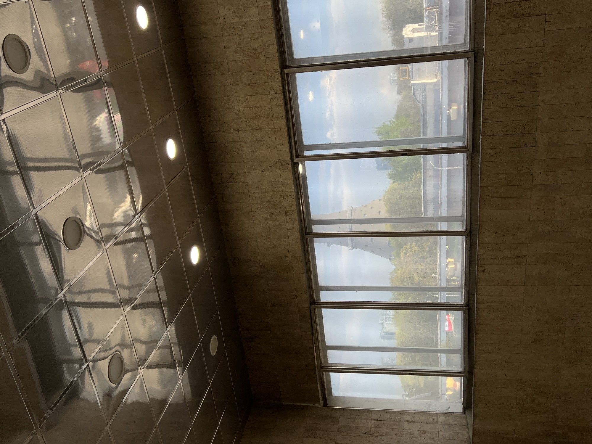The image shows an indoor view, likely a building interior in Khimki, Russia. The perspective is from below, looking upwards at a ceiling with recessed lighting and a set of tall, vertically stacked windows. The ceiling appears to be comprised of square or rectangular panels with a glossy, reflective surface. Circular recessed lights are evenly spaced throughout the ceiling. Through the windows, an external urban scene is visible, including a distant structure that might be a tower or monument. Trees and buildings are also discernible, suggesting a city landscape. The walls on either side of the windows are made of a textured, possibly stone or concrete material, providing a neutral backdrop to the scene.