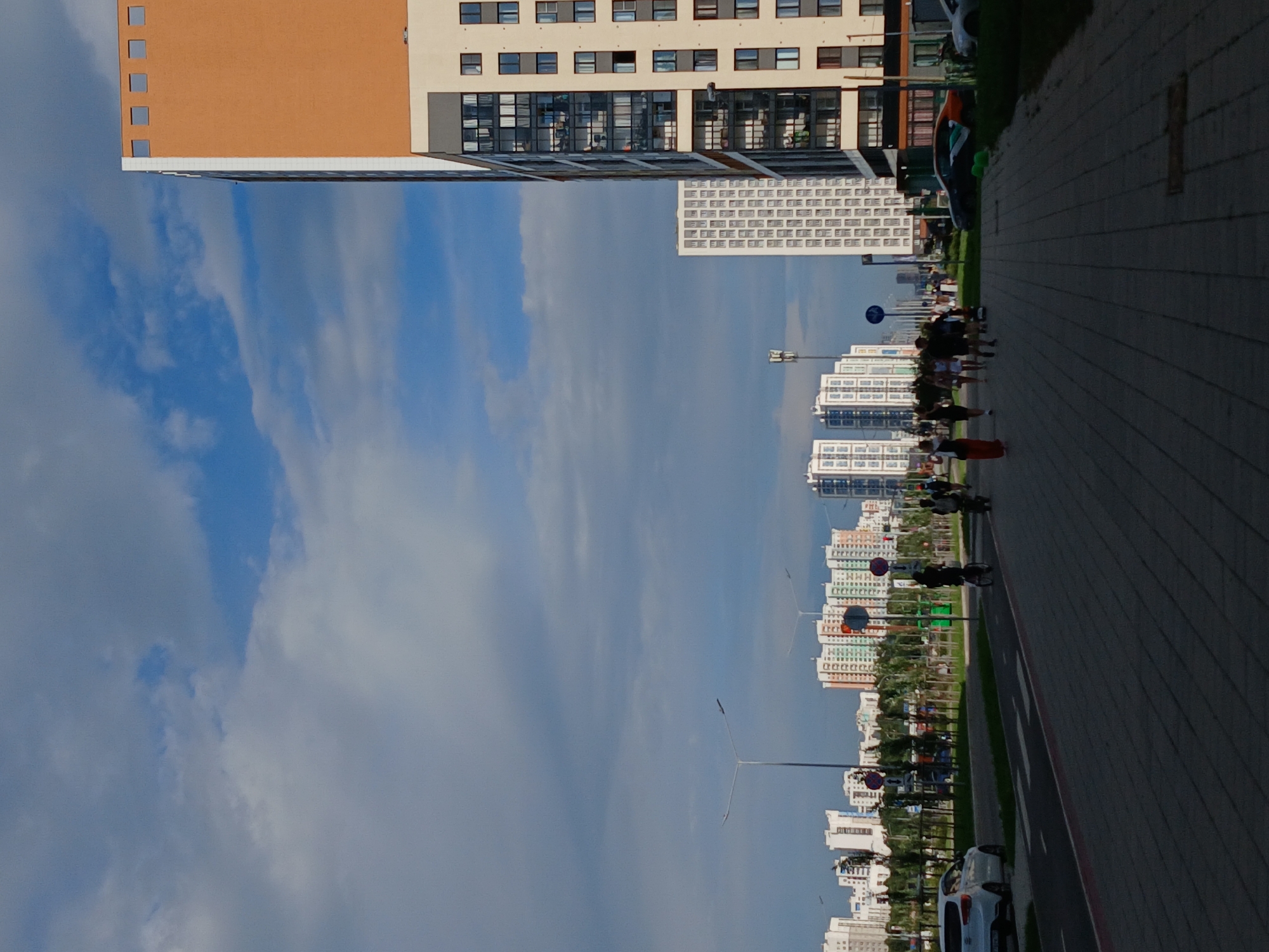 This image depicts a cityscape in Yekaterinburg, Russia, featuring a modern residential district under a cloudy sky. In the foreground, a paved pedestrian walkway runs parallel to a street. Several people are walking on the walkway, some individually and some in small groups. A person is riding a bicycle on the street. A white car is parked on the side of the street, and other vehicles are visible in the distance.
The middle ground is dominated by a row of multi-story apartment buildings, varying in architectural style and color. Some buildings have orange and white facades, while others are primarily white with dark window frames. Trees and landscaping line the street between the buildings and the walkway. Traffic lights and streetlights are also present.
The background reveals a dense collection of taller apartment buildings, extending towards the horizon. The sky is a mix of blue patches and dense gray clouds. The overall impression is that of a developing urban area with a focus on residential housing.