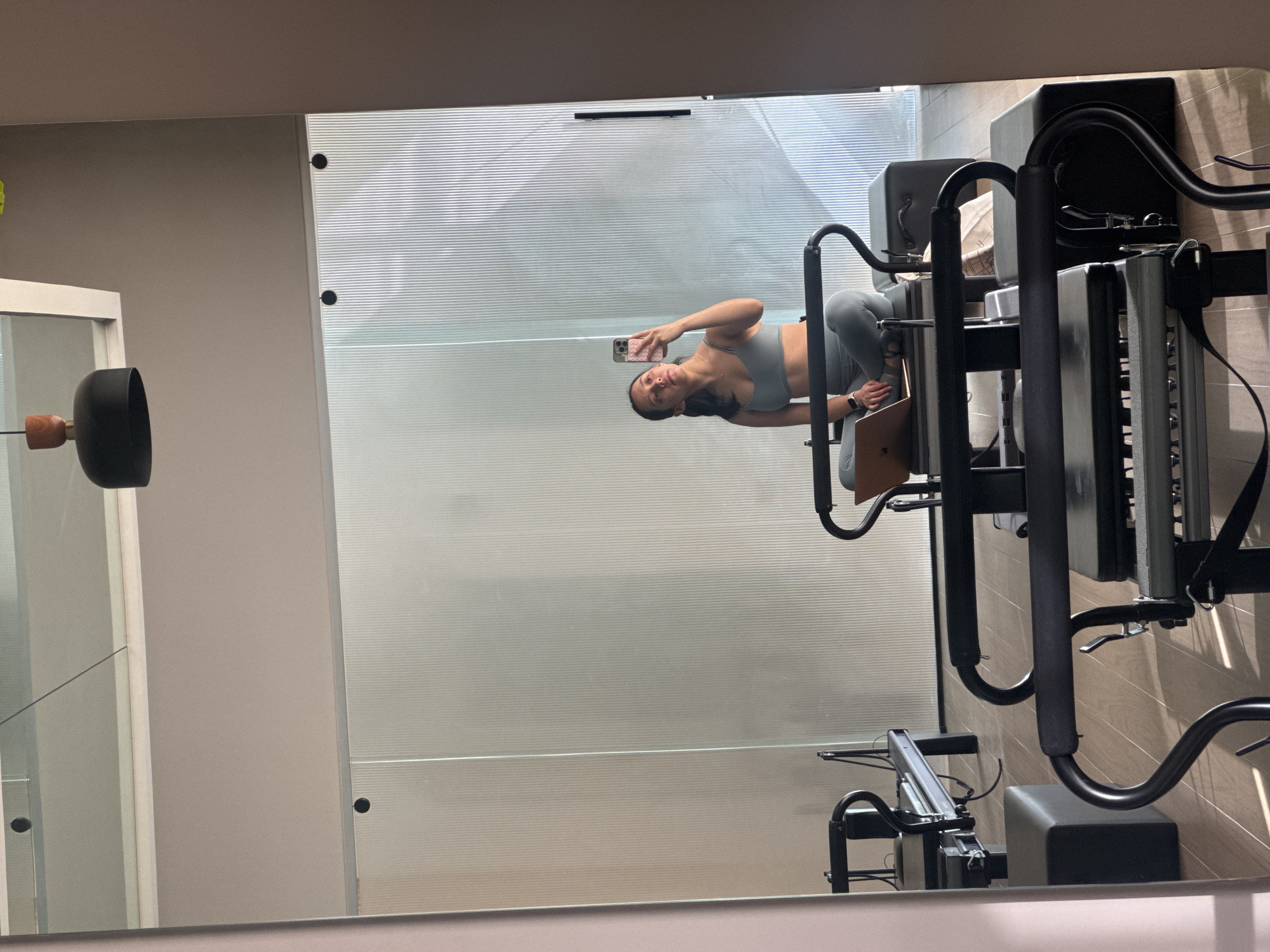 A young woman captures a mirror selfie in what appears to be a fitness or Pilates studio. She is positioned to the right of the frame in the reflection, wearing a light grey-blue sports bra and matching leggings. Her dark hair is pulled back, and she looks directly at the camera of her smartphone, which is held in her right hand and features a pink patterned case. A black smartwatch is visible on her left wrist.
To her right, a black and grey Pilates reformer machine is prominently featured, and a laptop is open on its platform next to her. The background behind the woman in the reflection consists of a wall of opaque, horizontally striped glass, suggesting a window or partition, allowing natural light into the space. The studio walls appear to be a light beige or grey. On the far left of the reflection, a decorative light fixture with a black dome and wooden accents is partially visible. The overall scene indicates daytime due to the ambient natural light. The woman maintains a neutral expression, gazing into the camera. No discernible text is visible on any objects or surfaces. The setting suggests a modern exercise facility in San Bernardino Tlaxcalancingo, Mexico.