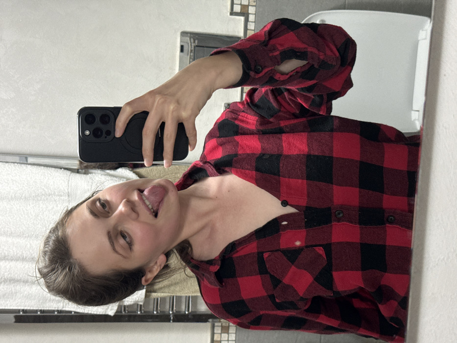 The scene depicts an interior bathroom located in Cava de' Tirreni, Italy. A young adult female is visible in a mirror reflection, capturing a selfie using a black smartphone held in her right hand. The smartphone features a multi-lens camera array and a circular attachment on its case. The female has light brown hair styled upwards and wears a red and black plaid flannel shirt, partially unbuttoned. Her head is tilted slightly to the viewer's left, her eyes are partially closed, and her tongue is extended out of her mouth.
The background, also visible in the mirror, shows a white wall with a metal towel rack holding a light-colored towel to the upper left. A horizontal strip of mosaic-patterned tiles (comprising brown, beige, and white tones) is visible at mid-height along the wall. To the lower right, a white toilet bowl and seat are reflected. A rectangular metallic fixture is mounted on the wall to the right of the toilet.