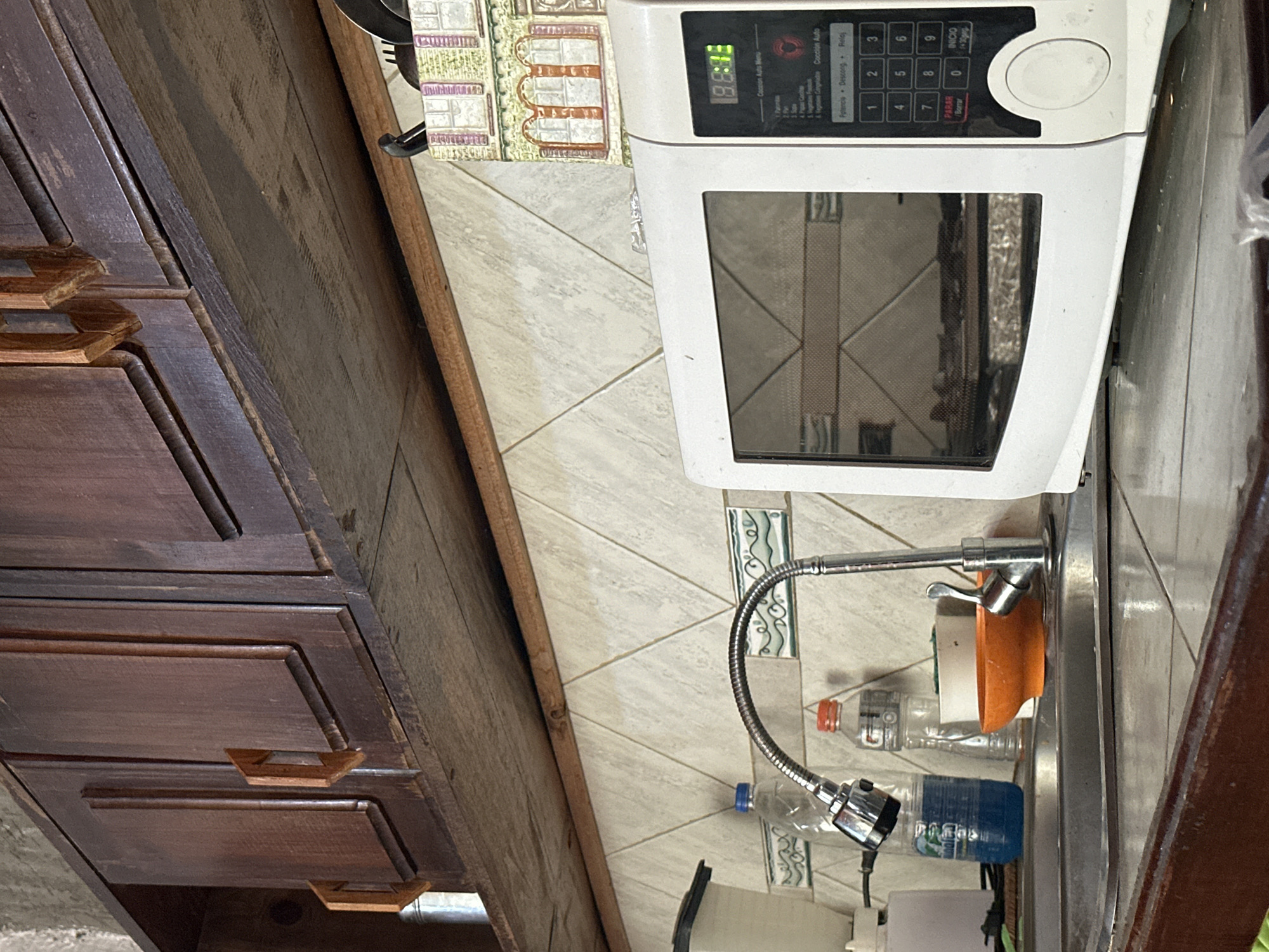 The image depicts an interior kitchen scene in Juan Griego, Venezuela. Dark brown wooden upper cabinets with carved wooden handles are mounted above a lighter wood shelf. Below the shelf, a backsplash features light-colored square tiles arranged diagonally, separated by dark grout lines. Interspersed within the backsplash are decorative rectangular tiles with a green/blue wave-like pattern. A stainless steel dual-basin sink is centered in the lower-left foreground, equipped with a flexible gooseneck faucet. To the left of the faucet, a blue plastic cup and a plastic bottle containing blue liquid are visible, along with a white soap dispenser. To the right of the faucet, another clear plastic bottle, an orange bowl, and a white container are present. On the countertop to the right of the sink, a white microwave oven is situated. Its digital display shows "18:83" in green, and its control panel is labeled in Spanish, including "Parar/Borrar" and "Inicio." Above and to the right of the microwave, a decorative utensil holder shaped like a building with windows and arches contains dark utensils. The countertop in the foreground is tiled and edged with dark wood. No people are visible, and no active interactions or actions are depicted.