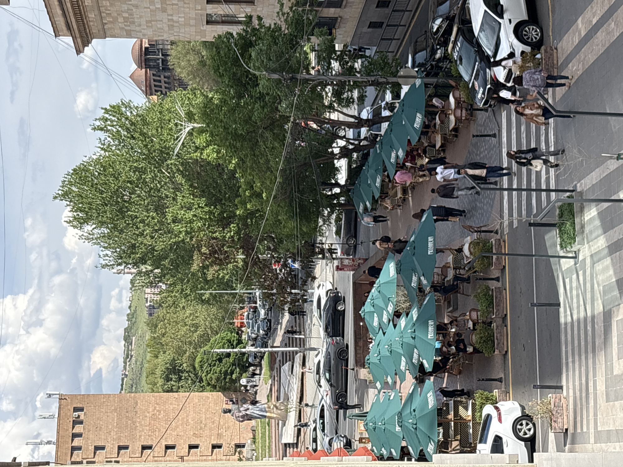 The image shows a street scene in Yerevan, Armenia. A row of green umbrellas shades outdoor seating for a restaurant along the sidewalk. Pedestrians are walking on a crosswalk. Cars are parked along the street and driving. A large tree dominates the scene. Buildings line the street, including a brick building in the foreground and a building with a dome visible in the distance.