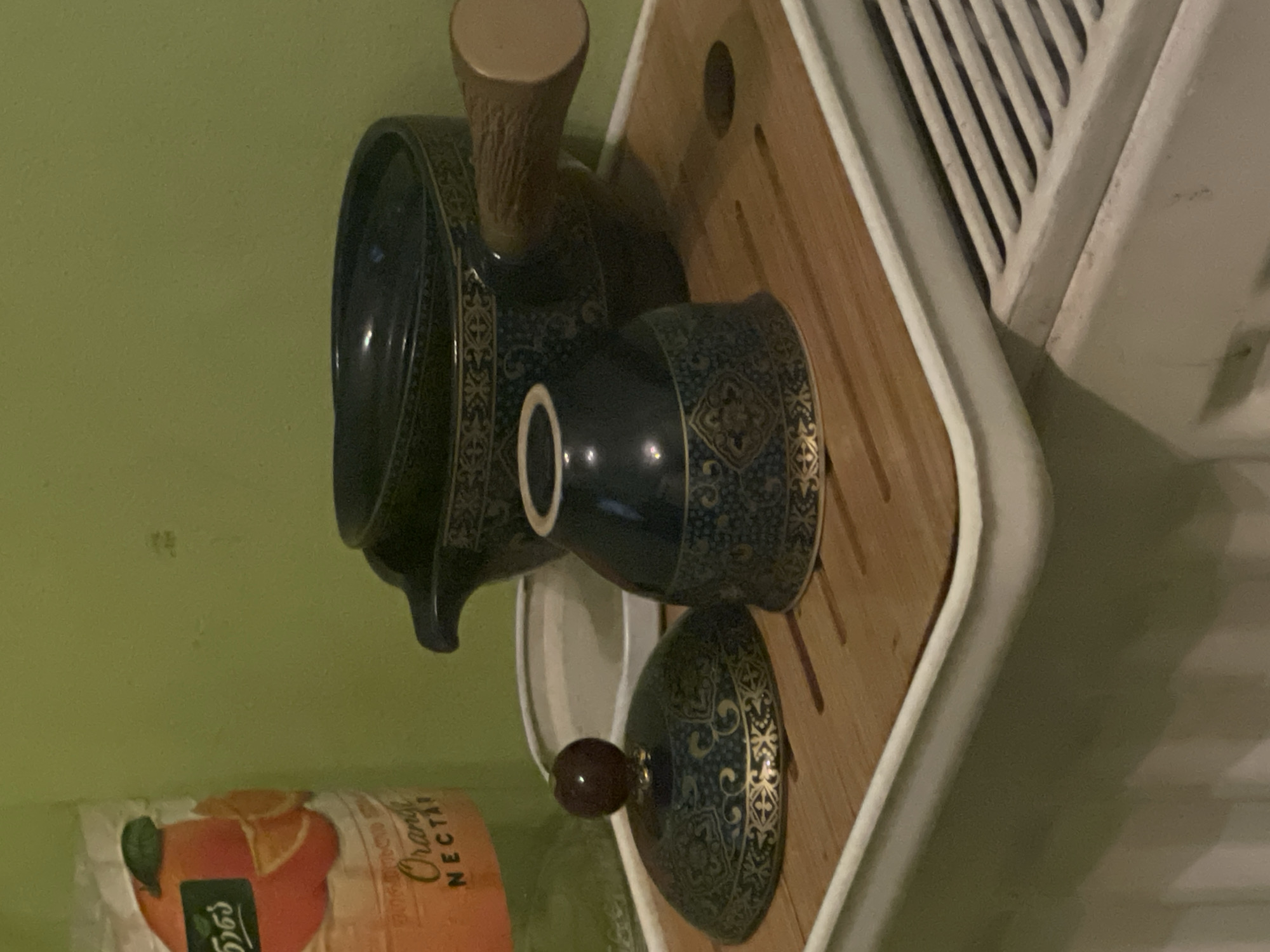 The image depicts a close-up of a dark blue, ornate tea set displayed on a bamboo tray. The tea set includes a teapot with a wooden handle, a lid with a small, dark knob, and a cup. The items are decorated with a gold, intricate pattern. To the left of the tea set, a portion of a bottle with an orange label featuring an illustration of oranges and text in Georgian script is visible. The background wall is painted a light green. The tea set rests on a white tray, which is positioned on a surface that appears to be a radiator or a similar heating element with horizontal slats. The scene suggests an indoor setting, possibly a kitchen or dining area, and the presence of the radiator indicates a cool environment. The overall impression is one of a still life arrangement, emphasizing the aesthetic details of the tea set and its surroundings. The location is identified as Batumi, Georgia.