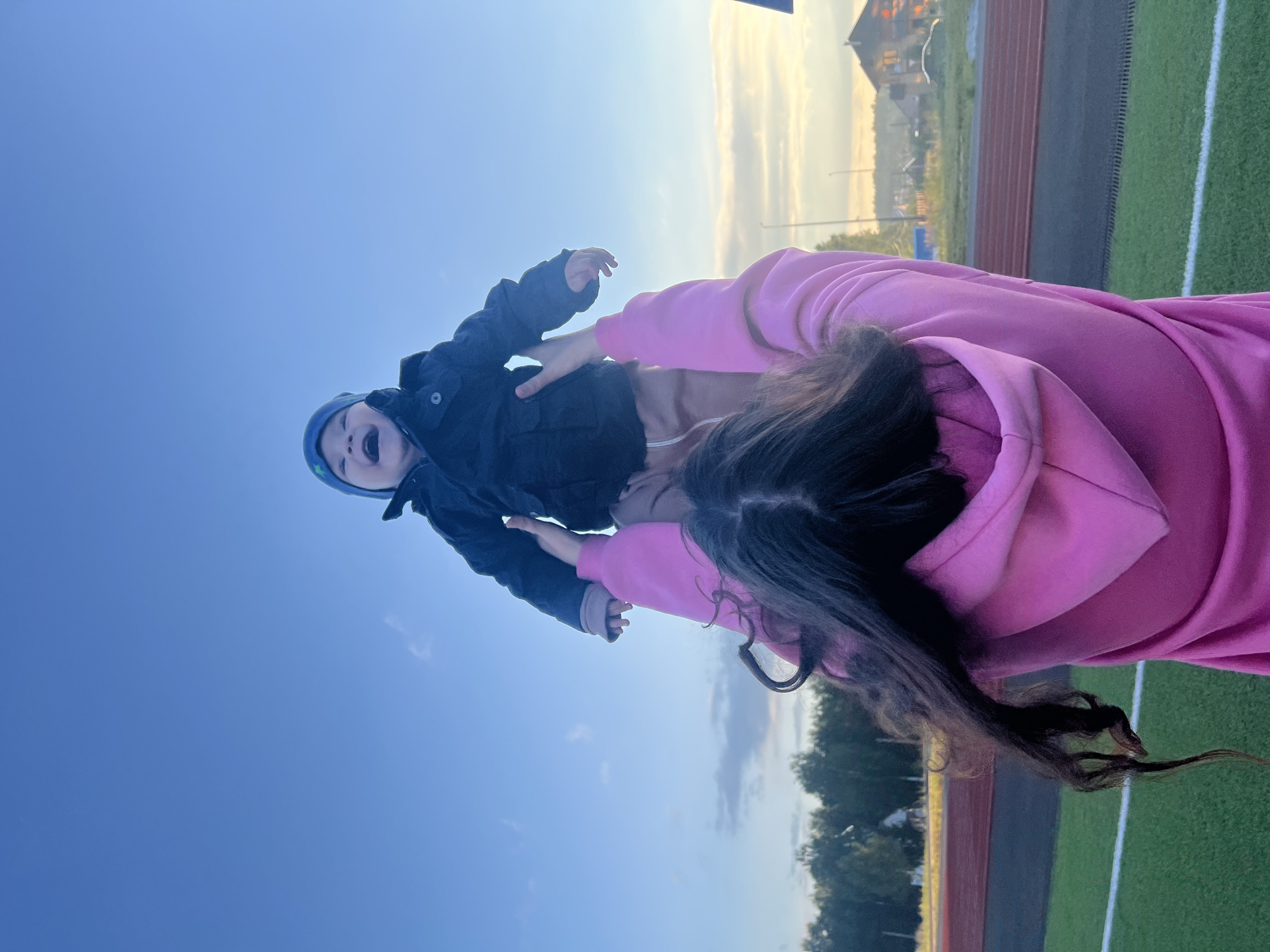 In Noviy, Russia, a woman in a pink hooded sweatshirt throws a baby up into the air against a clear blue sky. The baby, wearing a blue hat and a dark jacket, is captured mid-air with a wide, joyful expression. The woman's long, dark hair cascades around her shoulders. In the background, a sports field with green turf and red running tracks is visible, along with a line of trees and some houses in the distance. The lighting suggests it is either early morning or late afternoon.