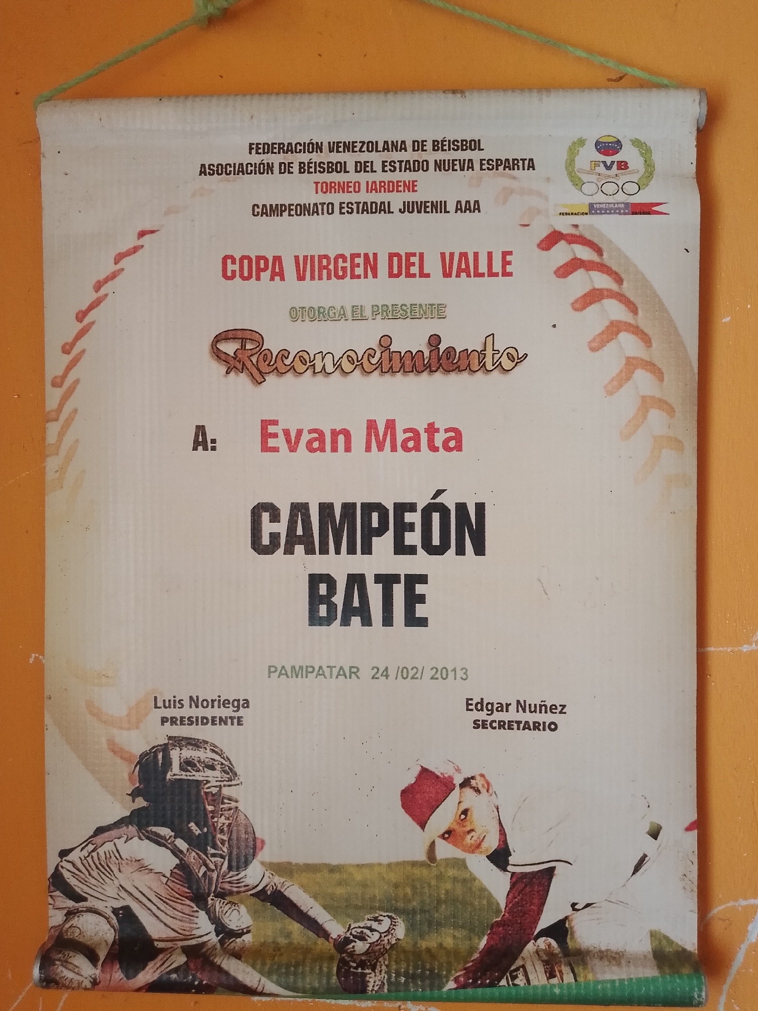 A rectangular paper or fabric banner, off-white with visible creases and light discoloration, hangs against a textured orange wall. A green cord is visible at the top, suspending the banner.
The banner is a recognition award for a baseball championship. At the top, the text reads: "FEDERACIÓN VENEZOLANA DE BÉISBOL" (Venezuelan Baseball Federation), "ASOCIACIÓN DE BÉISBOL DEL ESTADO NUEVA ESPARTA" (Baseball Association of Nueva Esparta State), "TORNEO IARDENE" (IARDENE Tournament), and "CAMPEONATO ESTADAL JUVENIL AAA" (State Youth AAA Championship).
In the upper right, a circular logo displays "FVB" (Federación Venezolana de Béisbol) within an olive wreath, above three Olympic-style rings. Below this, a small banner states "FEDERACIÓN VENEZOLANA DE BEISBOL".
The main title in large red letters is "COPA VIRGEN DEL VALLE" (Virgin of the Valley Cup). Below this, the phrase "OTORGA EL PRESENTE" (Awards the Present) leads to a stylized brown script reading "Reconocimiento" (Recognition).
The award is presented "A: Evan Mata", with the name in bold red letters. The achievement is specified as "CAMPEÓN BATE" (Batting Champion) in large black capital letters.
The document indicates "PAMPATAR 24 /02/ 2013" as the location and date of issuance. Pampatar is a city in Nueva Esparta state, Venezuela.
At the bottom, two signatories are listed: "Luis Noriega PRESIDENTE" (President) on the left, and "Edgar Nuñez SECRETARIO" (Secretary) on the right.
The banner's background incorporates stylized red baseball stitching patterns along its left and right edges. Two photographic images are placed at the bottom: on the left, a baseball catcher in full protective gear is in a crouched position; on the right, a young baseball player wearing a red cap and a white uniform is looking towards the viewer. Both images are in muted colors with a green field background.
