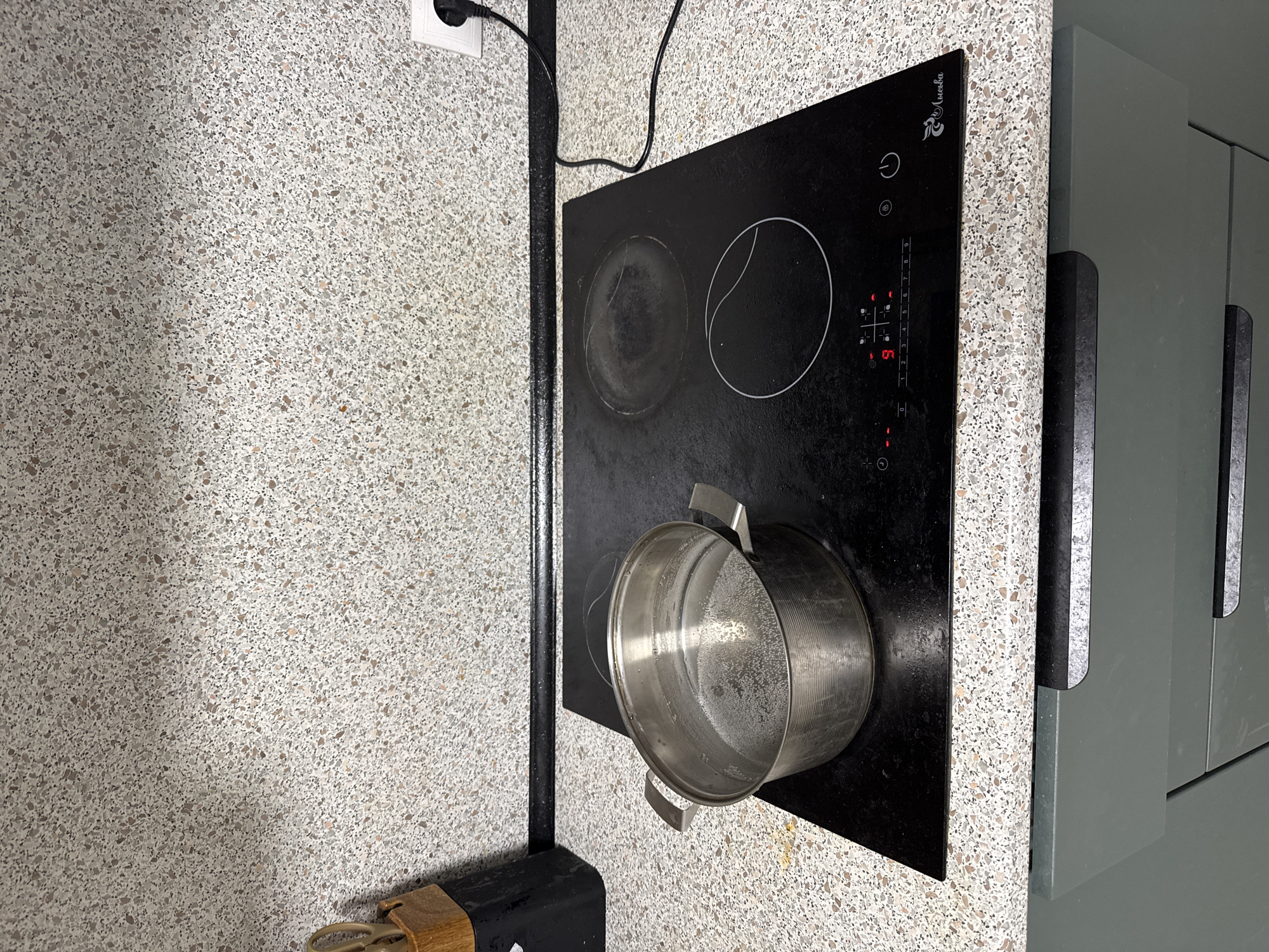 A top-down view shows a kitchen counter scene. A black glass cooktop with two visible circular heating zones sits to the right of a black metal vertical bar. A silver-colored pot is on the lower right corner of the cooktop, partially covering one heating zone. The cooktop's digital display shows the number "4". To the right of the cooktop are a series of green kitchen cabinets. The counter and wall behind the cooktop are covered with a speckled beige and gray material. A white electrical outlet with a black plug is visible on the wall above the vertical bar. The photograph provides no definitive evidence to confirm it was taken in Kazan, Russia.