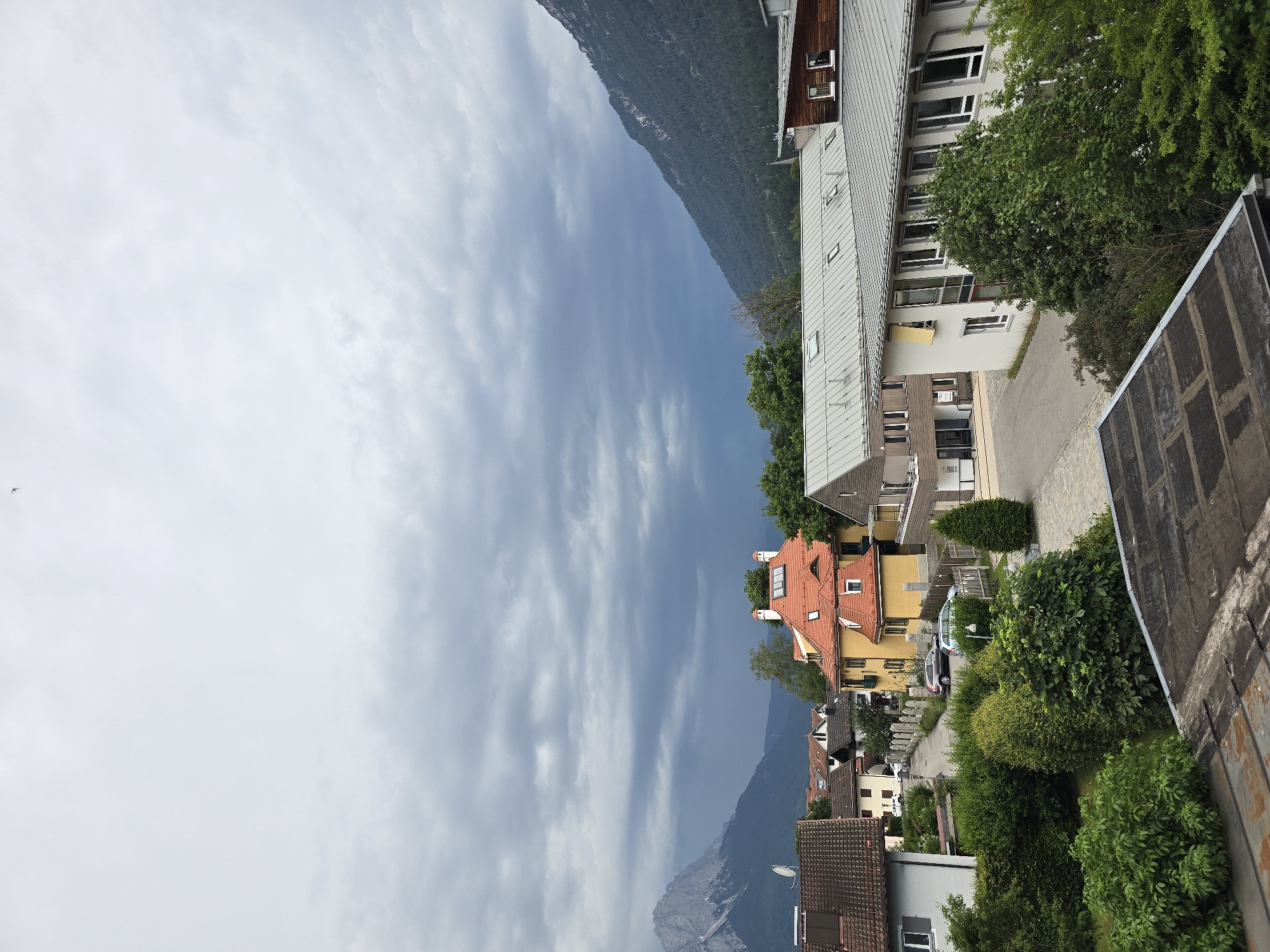 The image shows a view of the town of Garmisch-Partenkirchen, Germany, with buildings, trees, and mountains in the background. The sky is cloudy and overcast, with some darker clouds suggesting impending rain.
In the foreground, there are rooftops of buildings, some of which appear to be residential homes with tiled roofs. To the left, a white building with a dark tiled roof is visible. In the middle ground, several houses with bright orange or yellow roofs stand out. These houses have traditional architectural elements, such as dormer windows and chimneys. Adjacent to these houses is a modern-looking building with a light-colored facade and a large number of windows, suggesting it might be an office building or a school.
A street runs through the middle of the scene, with parked cars visible. The street is lined with fences and hedges. Lush green trees and bushes are abundant throughout the scene, providing a contrast to the built environment.
In the background, the imposing presence of the Alps mountains is evident. A snow-capped peak is visible on the left, and a densely forested mountain slopes down on the right. The weather appears to be changing, as indicated by the dark clouds gathering over the mountains. No people are visible in the image. The overall impression is a picturesque European town nestled against a dramatic mountain landscape under a moody sky.