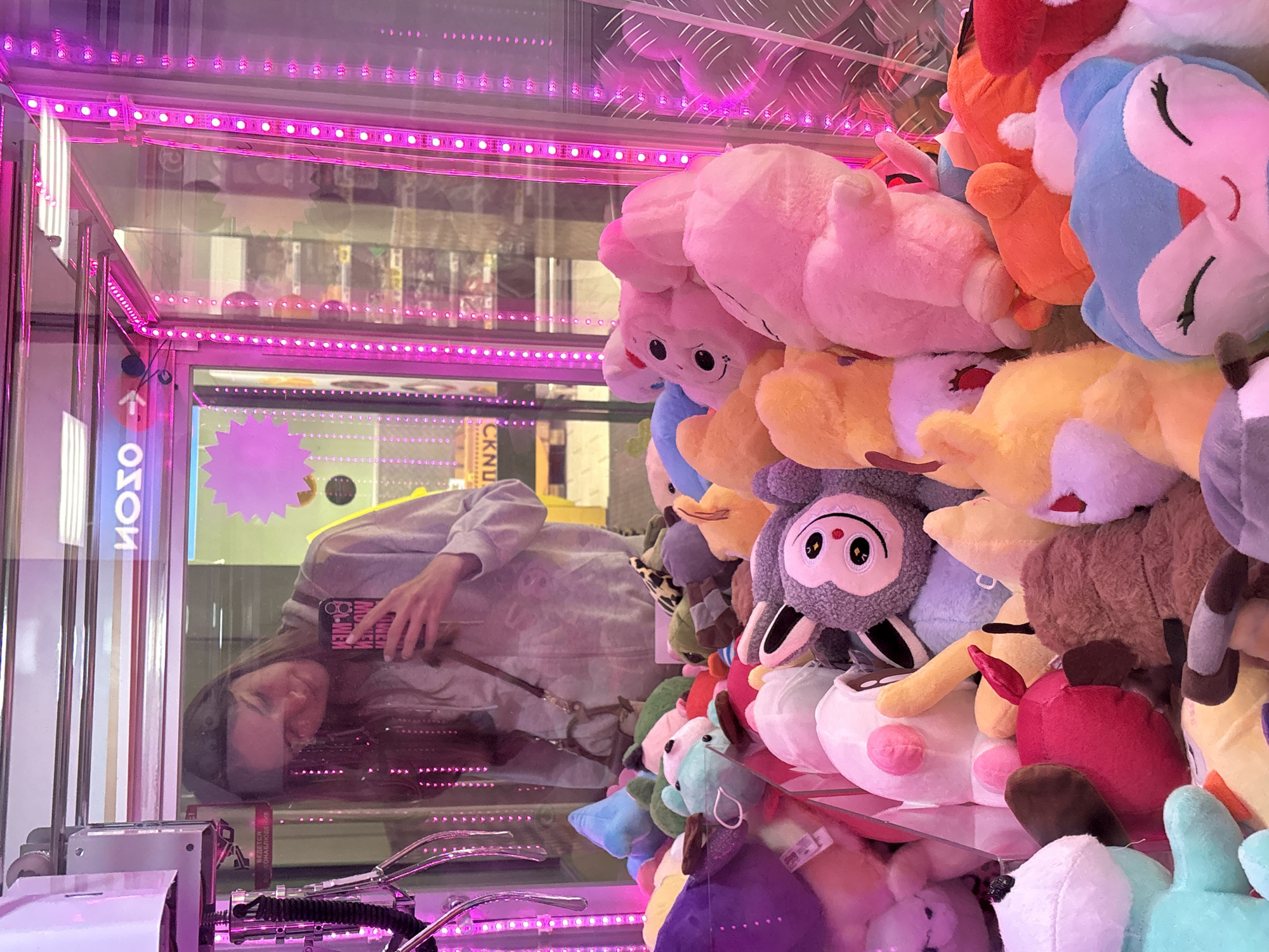 A person is visible in the reflection of a claw machine full of plush toys. The claw machine is illuminated with pink LED lights. The plush toys vary in color and include a pink toy, a yellow toy, a blue toy, and a grey toy. The person in the reflection is holding a phone. There is a sign in a foreign language, likely Russian, on the claw machine. The location is Perm, Russia.