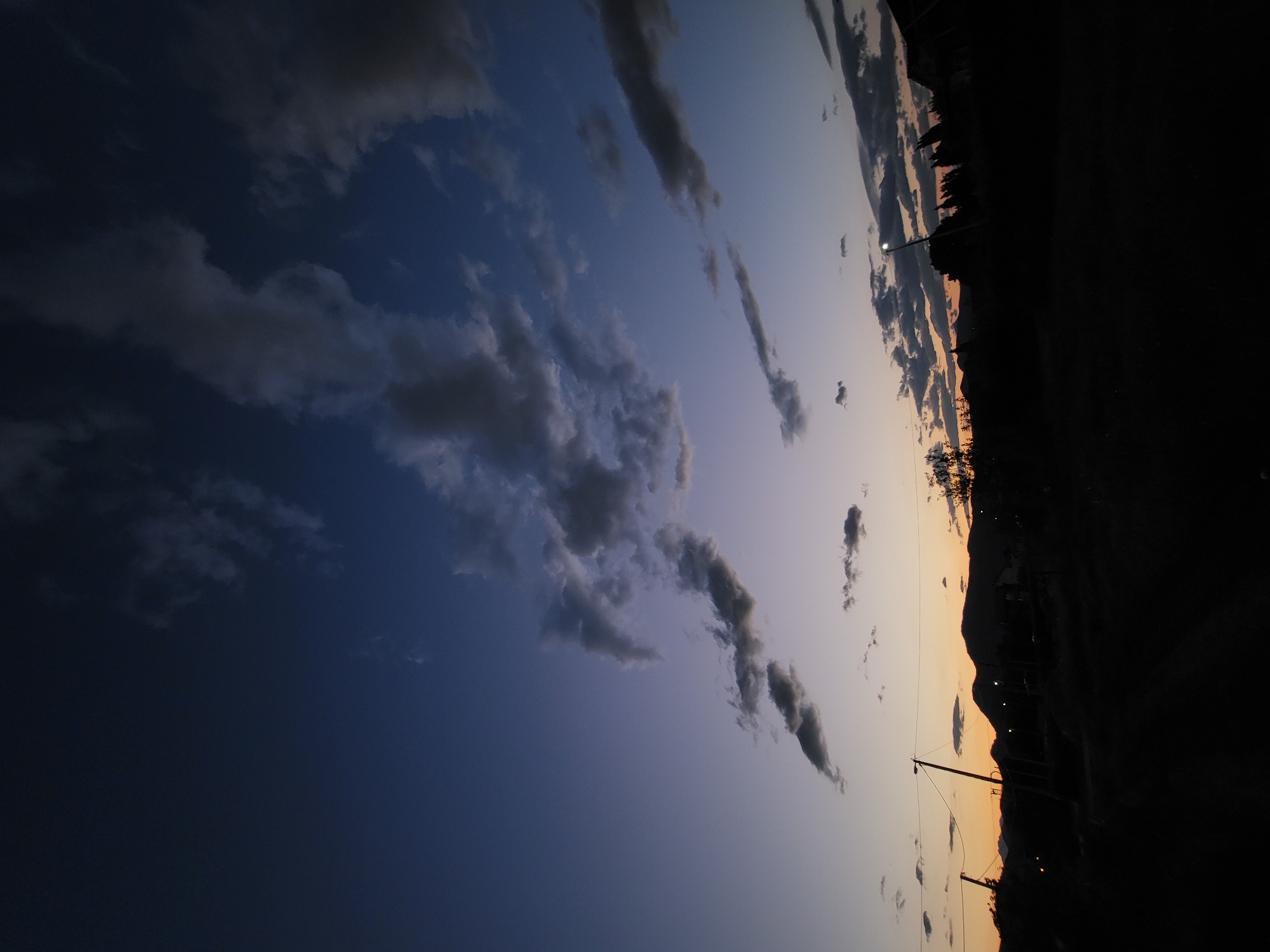 The image captures a serene twilight sky, presented in a portrait orientation. The sky displays a striking gradient, transitioning from deep indigo blue in the upper left to softer shades of purple, orange, and pale yellow towards the lower right. Numerous dark, wispy clouds are scattered across the sky, appearing as silhouettes against the lighter portions.

Along the right edge of the frame, the silhouette of a mountainous or hilly landscape defines the horizon. Below this, the foreground is shrouded in deep shadow, revealing indistinct shapes of trees, bushes, and possibly some structures. Several small, bright points of light, likely streetlights or lights from distant buildings, punctuate the darkness along the horizon line. A prominent utility pole, with wires extending from it, stands silhouetted against the sky in the lower-middle right section of the image.

The scene depicts a calm evening or early morning atmosphere. There are no people visible, nor any discernible text. The muted color palette and silhouetted elements create a tranquil and contemplative mood, showing a natural landscape transitioning between day and night in Mahala, Montenegro.