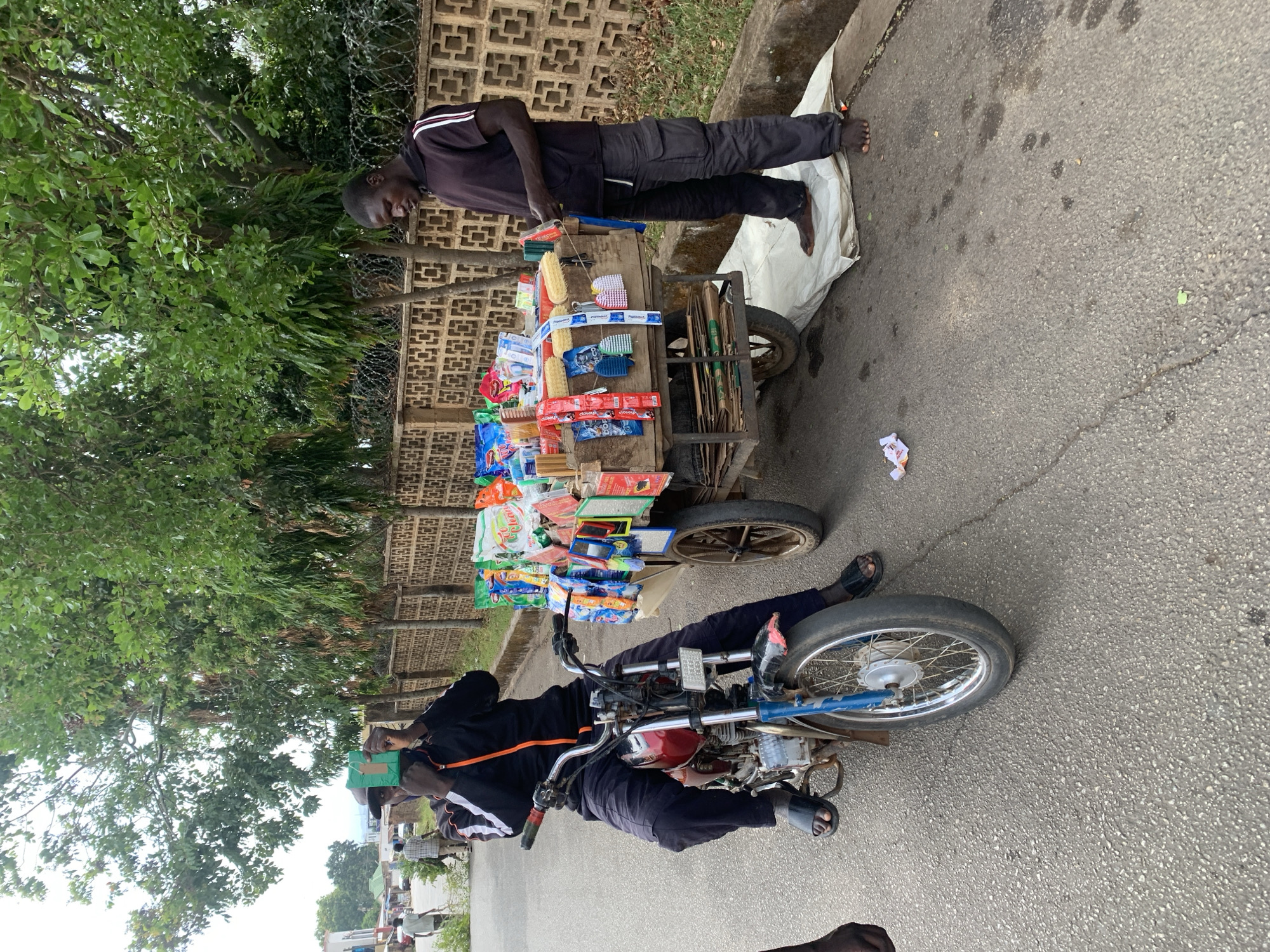 The image captures a vibrant street scene, likely in Jos, Nigeria, featuring two men and a vendor's cart. The main subject is a male street vendor, standing barefoot on a white cloth laid on the asphalt beside his two-wheeled wooden cart. He wears a black short-sleeved shirt with white stripes on the shoulders and dark trousers, looking towards the viewer with a slight smile.
His cart is heavily laden with a variety of goods, primarily household items and toiletries. Visible products include different types of scrubbing brushes (some corn-shaped, others with plastic handles), sponges, and numerous sachets and small packages of consumer goods. Brands such as "Close-up" (toothpaste sachets), "Mamee" (snack), and "Chana" are discernible on some packaging. Below the main display, stacked items resembling small plastic chairs or stools are visible.
To the left of the cart, another man is seated on a red and blue motorcycle, positioned partially behind the cart. He is wearing a dark long-sleeved top with an orange stripe down the arm and dark trousers, with black sandals on his feet. A green box or item is held in his left hand.
The background features a tall, light brown concrete block wall with a patterned design, topped with barbed wire. Lush green trees and dense foliage grow behind the wall, suggesting a verdant environment. The ground is asphalt, showing some wear and scattered debris. The overall impression is that of a bustling daytime market or roadside vending activity.