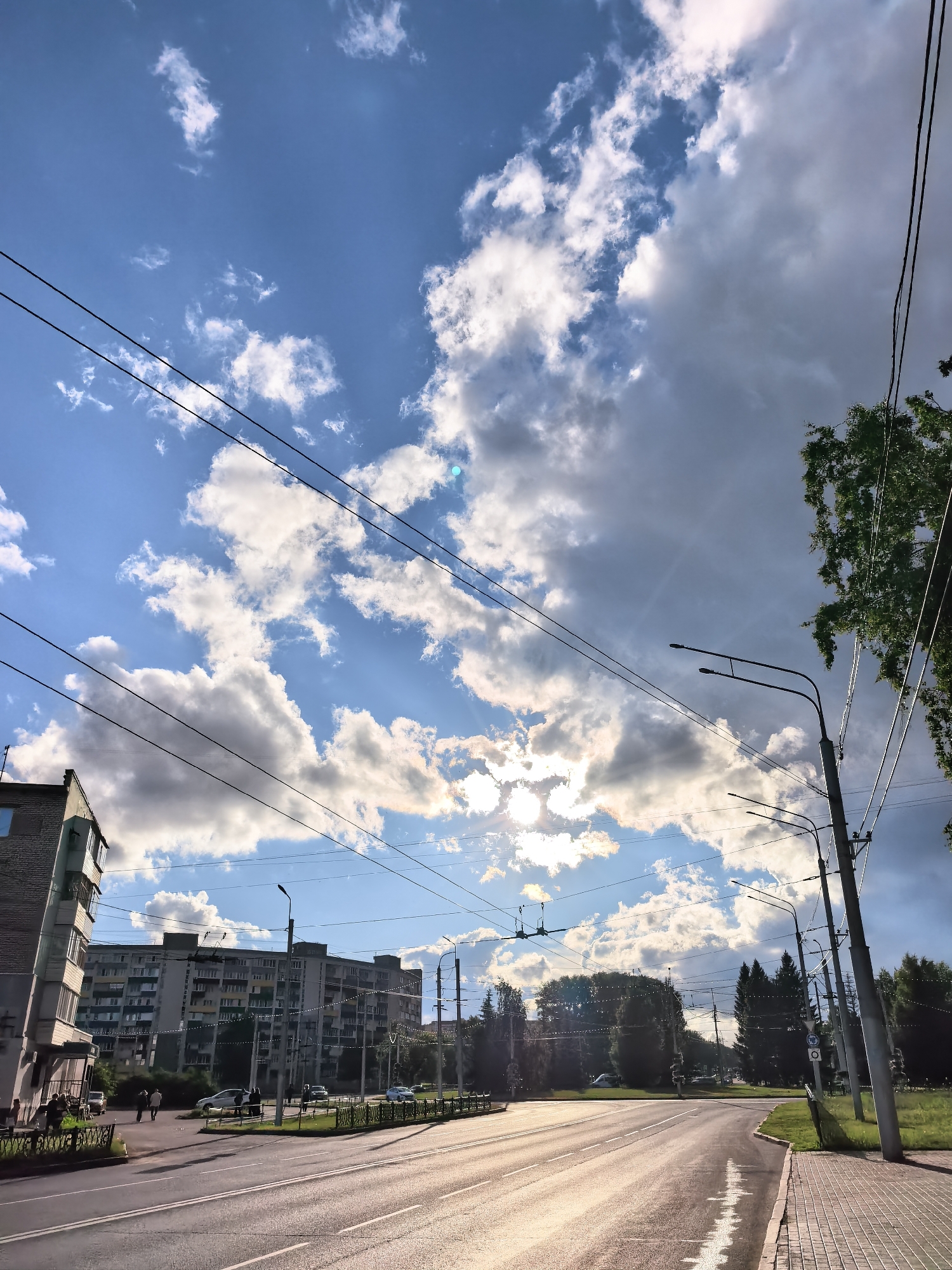 The image depicts a street scene in Sterlitamak, Russia, under a cloudy sky. A multi-story residential building is visible on the left, and a road stretches into the distance. Pedestrians are walking on the sidewalk, and cars are on the road. Overhead power lines cross the sky. Streetlights line the road. Trees are located on the side of the road.