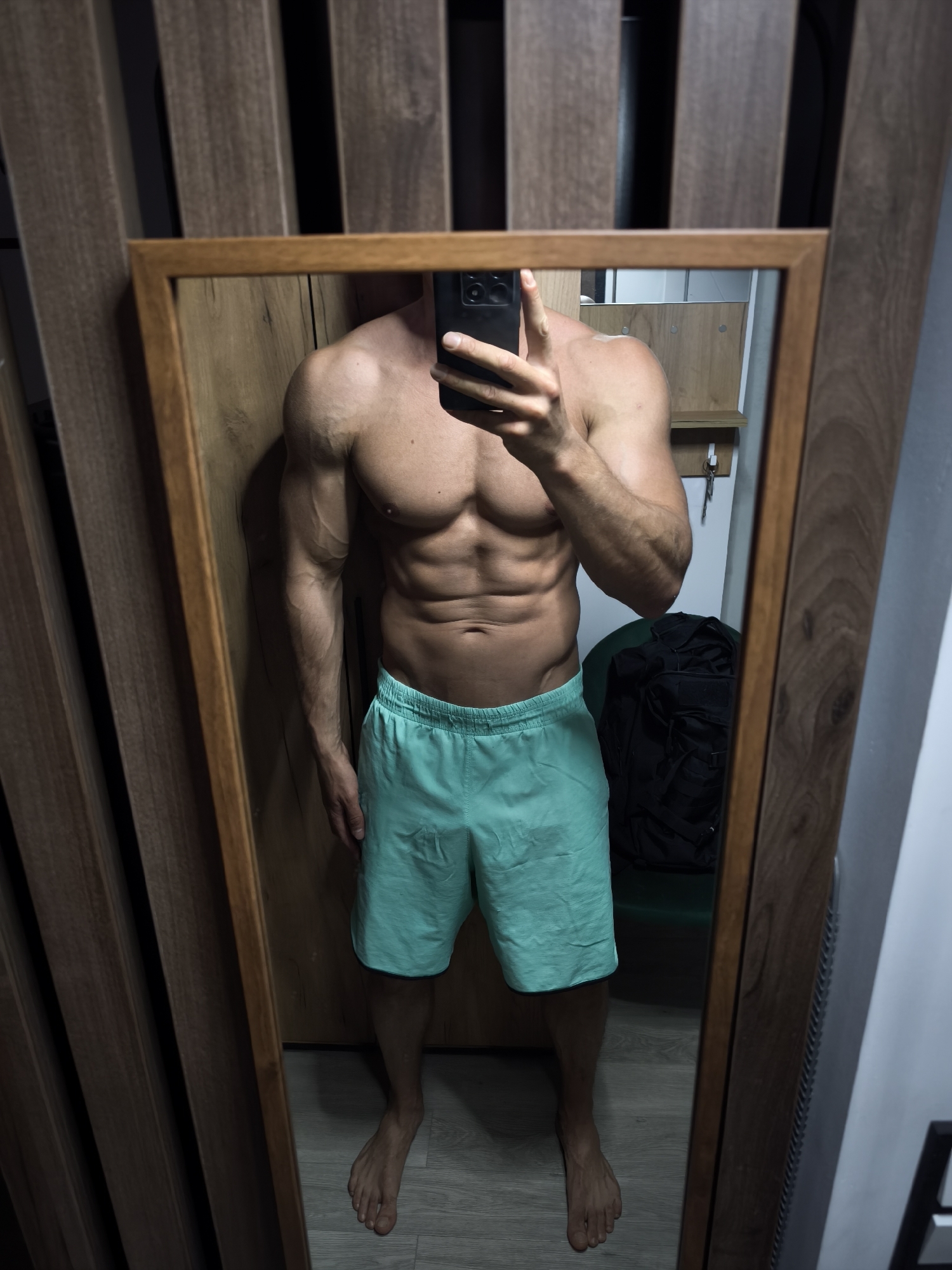 A shirtless, muscular man is taking a selfie in a mirror. He is wearing light blue shorts. The man's face is obscured by a black mobile phone. The mirror is framed in wood and reflects an interior with wooden paneling, a green chair, and a black backpack. The environment appears to be a room with a wooden feature wall to the left of the mirror. It's not possible to determine the location in Moscow, Russia, from the image.