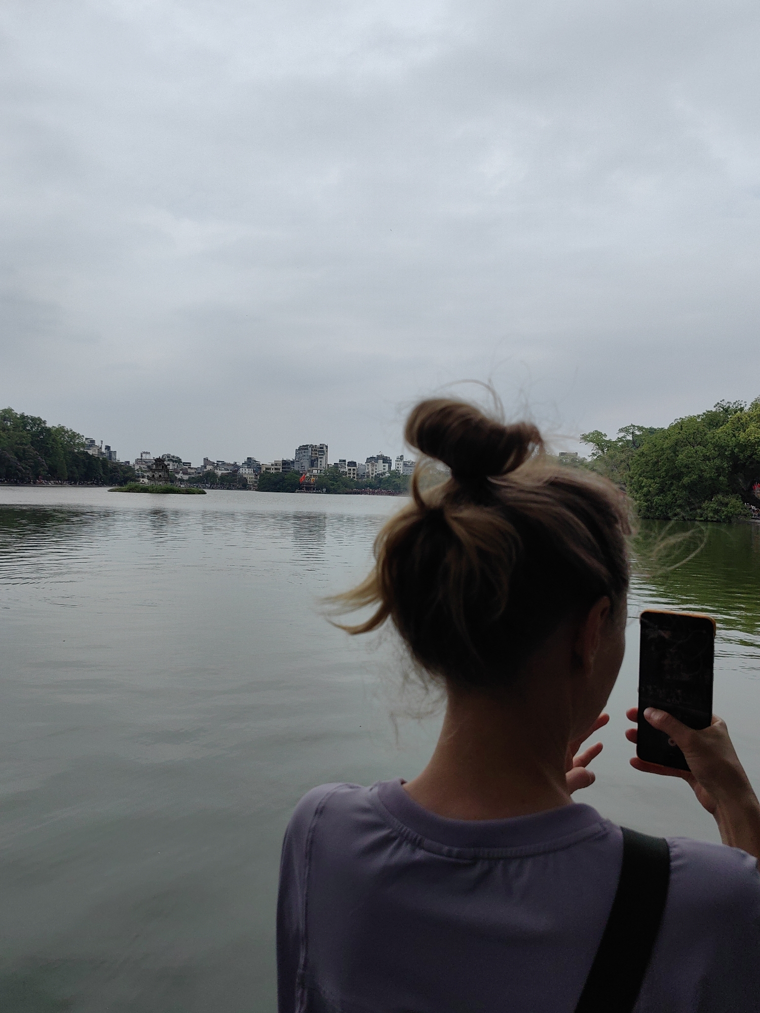 A woman is taking a picture with a smartphone of Hoàn Kiếm Lake in Hà Nội, Vietnam. The woman has brown hair styled in a bun. She is wearing a light purple shirt and a black strap is visible over her shoulder. In the background, the lake is calm with the city skyline visible on the opposite shore. An island with a pagoda is visible in the center of the lake. The sky is overcast.