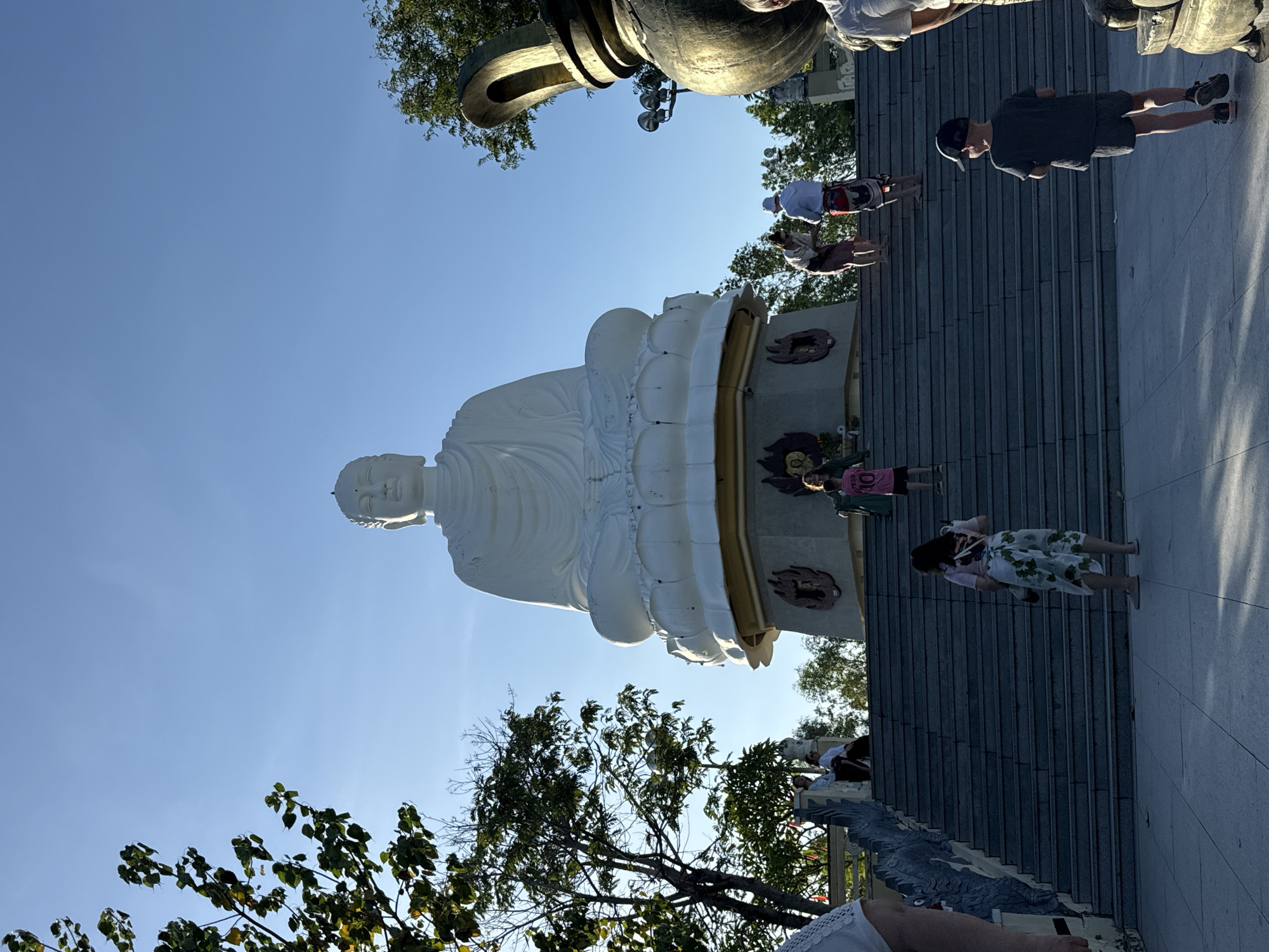 The image shows a large, white Buddha statue in Tay Nha Trang Ward, Vietnam. The statue is seated on a lotus flower platform, viewed from a low angle, against a clear blue sky. Several people are present, including children, walking or standing on a large staircase to the right of the statue. Green trees surround the base of the statue. Above, there is part of a large dark bell structure.