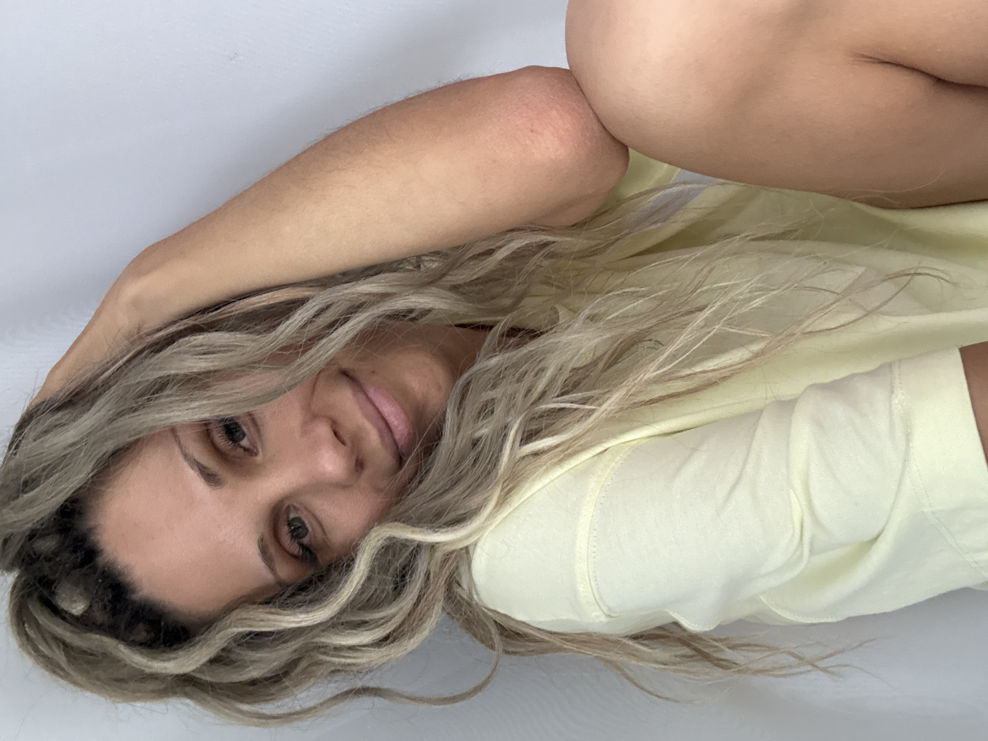 A woman with blonde, wavy hair pulled up into a ponytail and wearing a pale yellow top is captured in a close-up, head-and-shoulders shot. She is leaning her head against her arm, with her hand resting on the back of her head, and her gaze is directed towards the camera with a slight smile. Her left arm is bent at the elbow, with the elbow visible and slightly reddish. The background consists of a plain white wall. The image does not contain any information that would indicate a specific location in Rusanovo, Russia.