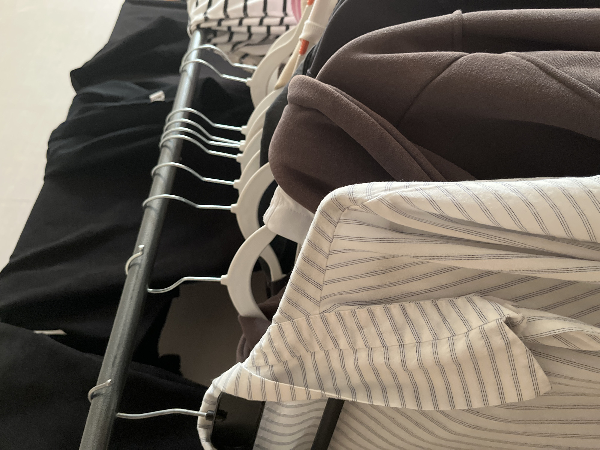 A close-up shot shows several garments hanging on a metal clothing rack. In the foreground, a white button-down shirt with thin blue vertical pinstripes is displayed prominently. To its right, a brown hooded sweatshirt is visible. Behind these, other dark-colored clothing items and a black garment are hanging. The lighting suggests an indoor environment, possibly a closet or a clothing store. There are no people or animals present in the image.