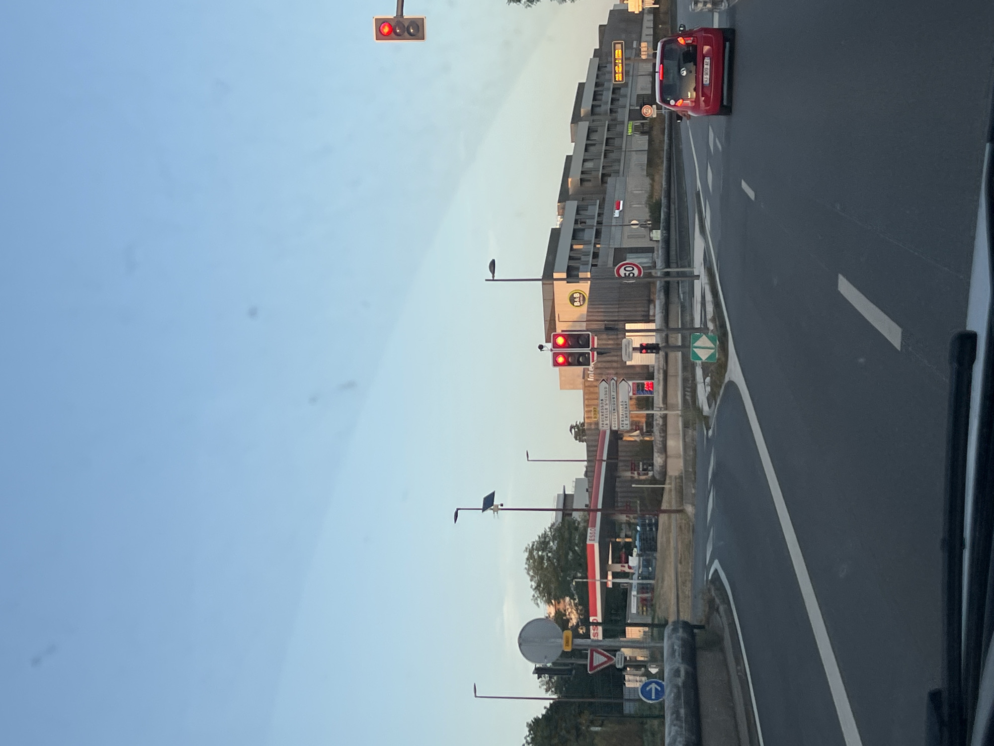 The image depicts a street scene in Le Chesnay-Rocquencourt, France, likely near a commercial area. In the foreground, a two-lane road is visible with white lane markings and a pedestrian crossing. On the right side of the road, a red car is driving away from the camera. In the middle ground, a gas station with red awnings is present, identified by "ESSO" signage. Adjacent to the gas station is a multi-story building with a sign for "B&B HOTELS." Traffic lights are visible, with one displaying red. Several road signs are present, including a directional sign with place names such as "Puteaux" and "Versailles." The sky is clear and blue, suggesting daytime. The scene is likely captured from a vehicle.