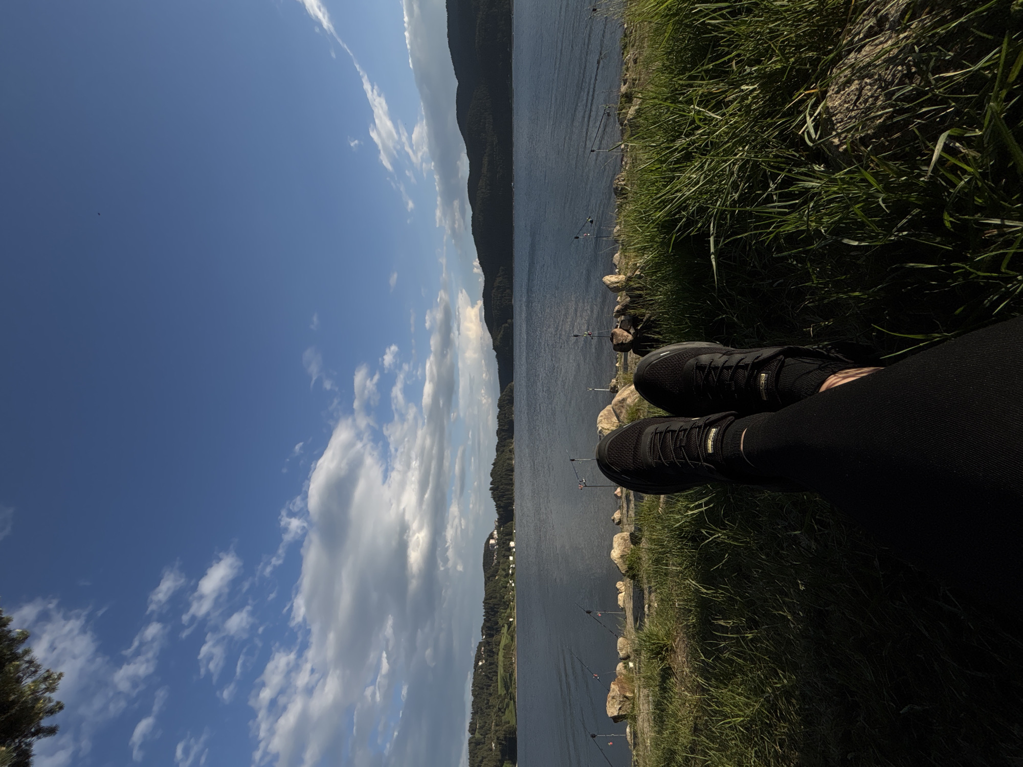 The image shows a first-person perspective of someone sitting at the edge of a lake or large body of water. The person is wearing black shoes and black leggings. To the right, there's grassy vegetation and rocky terrain. The water stretches out towards a tree-covered mountain range in the distance. Fishing rods are visible at the edge of the lake, suggesting fishing activity. The sky is predominantly blue with scattered clouds. The location is Unknown, Unknown.