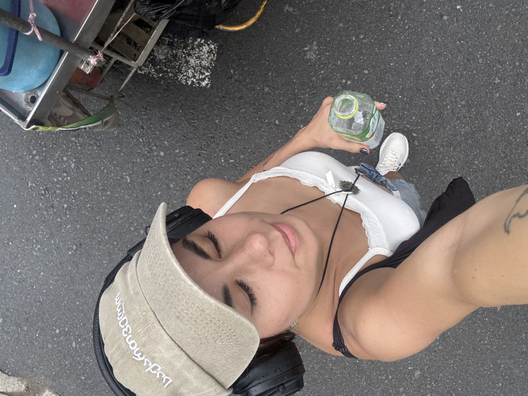 A woman wearing a light brown baseball cap with white writing, black headphones, a white lace-trimmed tank top, denim shorts, and white sneakers is holding a clear plastic bottle of green liquid. She is standing on an asphalt surface, likely a street or sidewalk. In the background, a metal cart with a blue bucket and pink twine is partially visible. The scene suggests an outdoor urban environment. The text on the cap is in Georgian script.