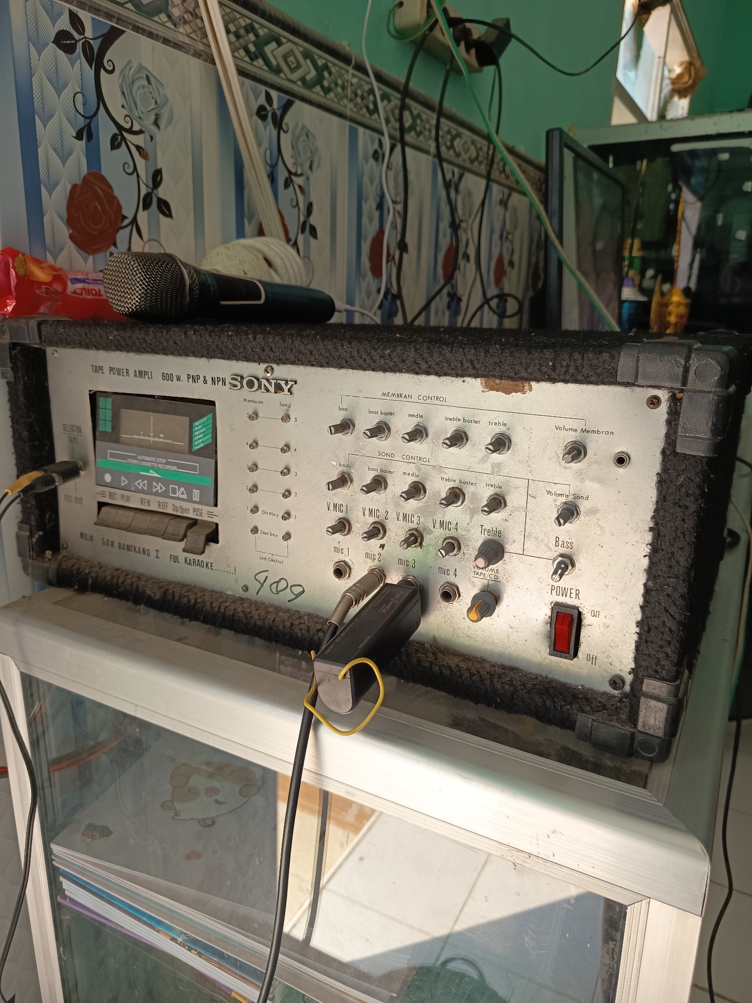The image shows a Sony Tape Power Amplifier, model 600 W, PNP & NPN, placed on top of a white cabinet with glass doors. The amplifier has a cassette player with various controls, including volume, bass, treble, and microphone inputs labeled V.MIC 1, V.MIC 2, V.MIC 3, and V.MIC 4. There is also a power switch labeled "on" and "off". Two microphones are resting on top of the amplifier. The amplifier and surrounding area appear dusty and aged, suggesting it is an older piece of equipment. The background shows a wall with a floral patterned wallpaper. The overall scene appears to be indoors, possibly in a residential setting. No individuals are present in the image. There is no visible indication that this location is specifically in Widoro, Indonesia.
