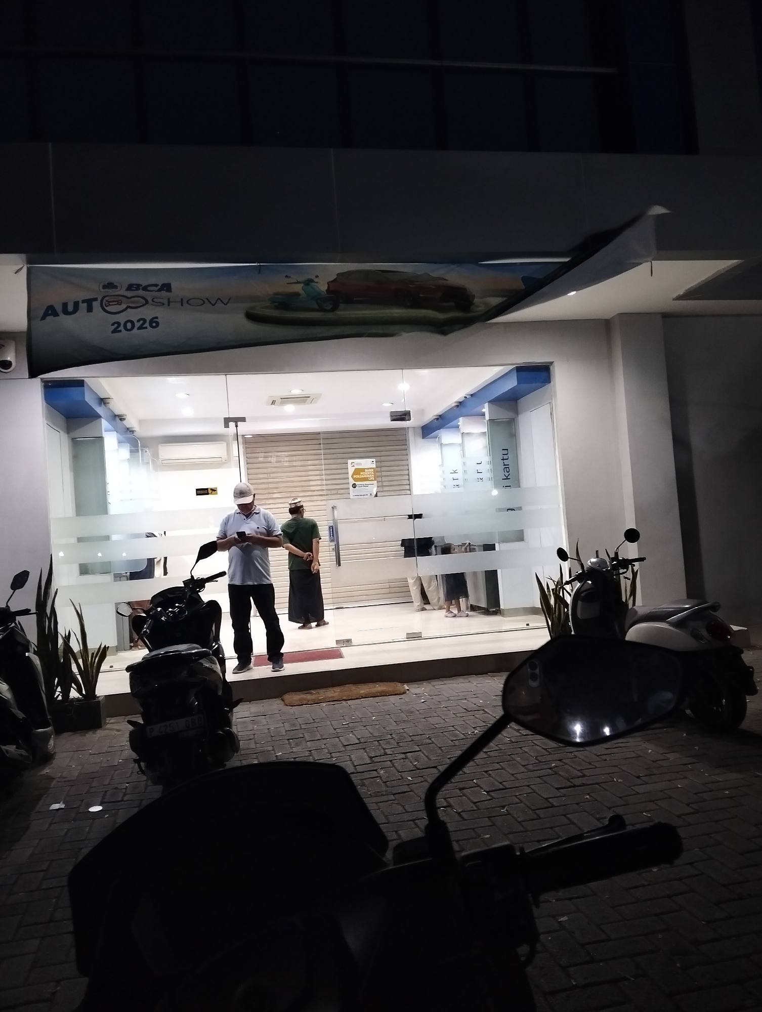 A nighttime exterior shot captures a BCA Auto Show banner with "2026" prominently displayed above the entrance of a building. The banner depicts a blue scooter and a red car. Two men stand outside a glass-fronted building. One man, wearing a white cap and light blue polo shirt, is looking at his mobile phone while standing next to a parked black scooter. A second man, wearing a green shirt and a traditional cap, stands further back near the entrance, looking towards the building. A third person, partially visible, stands in the doorway. Parked scooters are in the foreground and to the left of the building. The pavement is made of interlocking bricks. The scene appears to be in Muncar, Indonesia, likely a commercial area with the BCA bank visible through the glass.