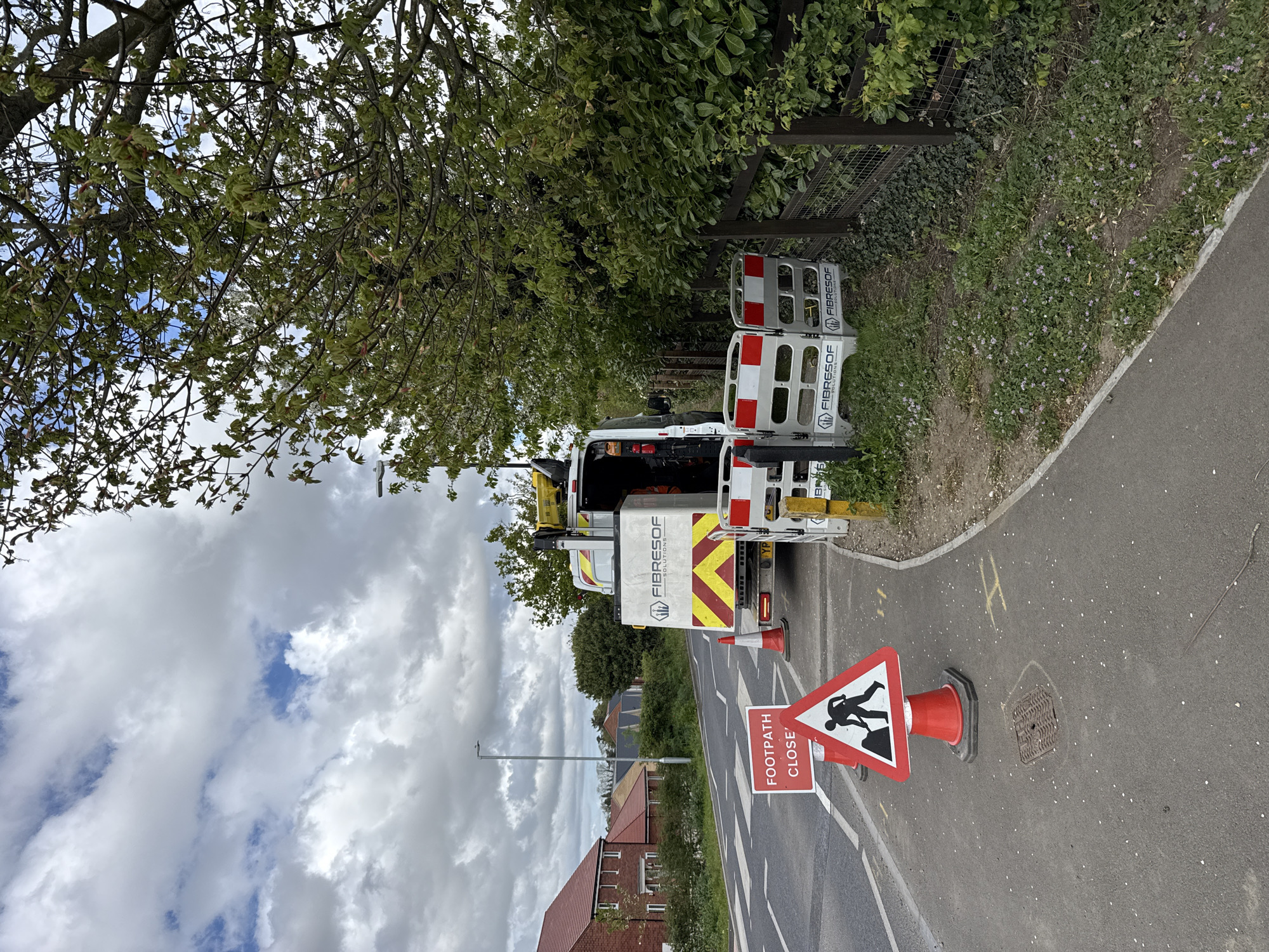 The image depicts a roadside scene in Mid Suffolk, United Kingdom. A white van, labeled "FIBRESOF SOLUTIONS", is parked partially on the paved sidewalk and the adjacent road. The rear doors of the van are open, revealing equipment inside. Two red and white barrier sections, also labeled "FIBRESOF", are positioned behind the van. A traffic cone with a "FOOTPATH CLOSED" sign is located in the roadway, near a manhole cover. The road has double white lines. A residential area, including brick houses, is visible in the background. A tree overhangs the van and the sidewalk. The sky is cloudy with patches of blue.