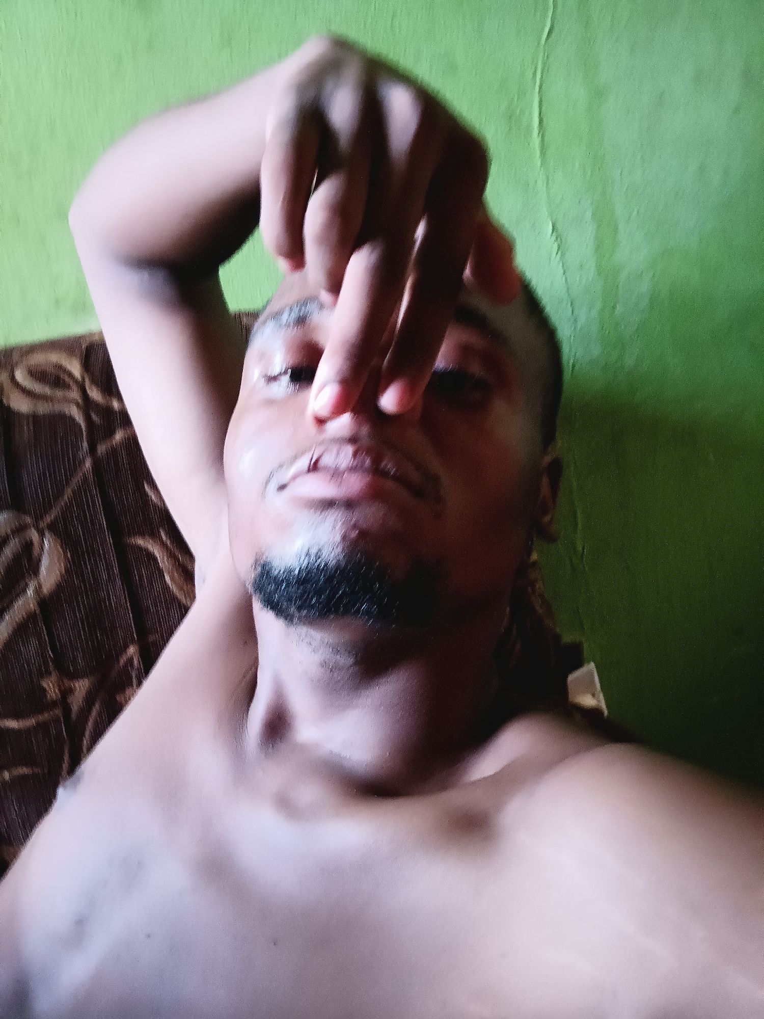 An indoor, close-up, upward-angle shot captures a young adult male, likely of African descent, from the chest up. He is shirtless, displaying his bare chest and shoulders. His right hand is positioned over his nose and forehead, with fingers spread, obscuring parts of his face. His face has a subtle sheen, possibly from sweat. He has a short beard and very short hair. His expression is mostly obscured but appears relaxed.
The background on the right and top consists of a vibrant, light green wall, showing some texture and minor imperfections, such as slight peeling or unevenness. On the left and bottom, a brown surface with a dark, intricate pattern is visible, suggesting the upholstery of a couch or chair. The overall lighting appears to be from an indoor source, casting some soft shadows. The image has a slightly blurry quality, particularly around the edges and background. No text is visible. The scene suggests a casual, personal moment within an indoor setting in Narayi, Nigeria.