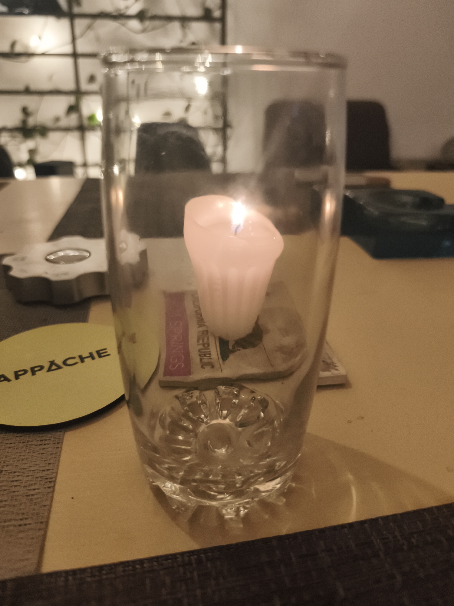 A clear glass cylinder encases a small, lit, white candle resting on a coaster, which is in turn on top of what seems to be a small map of Palm Springs in California. The glass sits on a table with a yellow and brown surface. A round yellow coaster with the word "APPACHE" printed on it is adjacent to the glass. To the left, a decorative metal object resembling a gear is partially visible. In the background, there is a table, chairs, and a wall with decorative lighting and plants. The image was taken in Mexico City, Mexico.