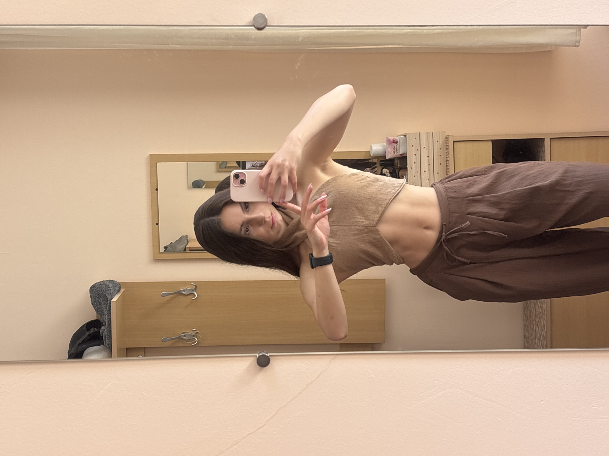 A young woman with dark hair stands in front of a full-length mirror, taking a selfie with her pink smartphone. She is wearing a cropped, halter-neck top in a light brown color and wide-legged, brown pants cinched at the waist with a drawstring. A black smartwatch is visible on her left wrist.
The background suggests an indoor setting, possibly a dressing area or entryway. To the left of the mirror is a tall, wooden panel with two silver coat hooks. Above the hooks, a shelf holds a grey knit hat and a dark bag. To the right of the woman, a wooden cabinet with drawers is partially visible, topped with various items including bottles of perfume and decorative small hearts. A second, smaller mirror is visible in the reflection on the cabinet. The walls are painted a pale peachy-pink color. The overall impression is that of a casual, personal space. The specific location within Varna, Bulgaria cannot be determined from the visual information alone.
