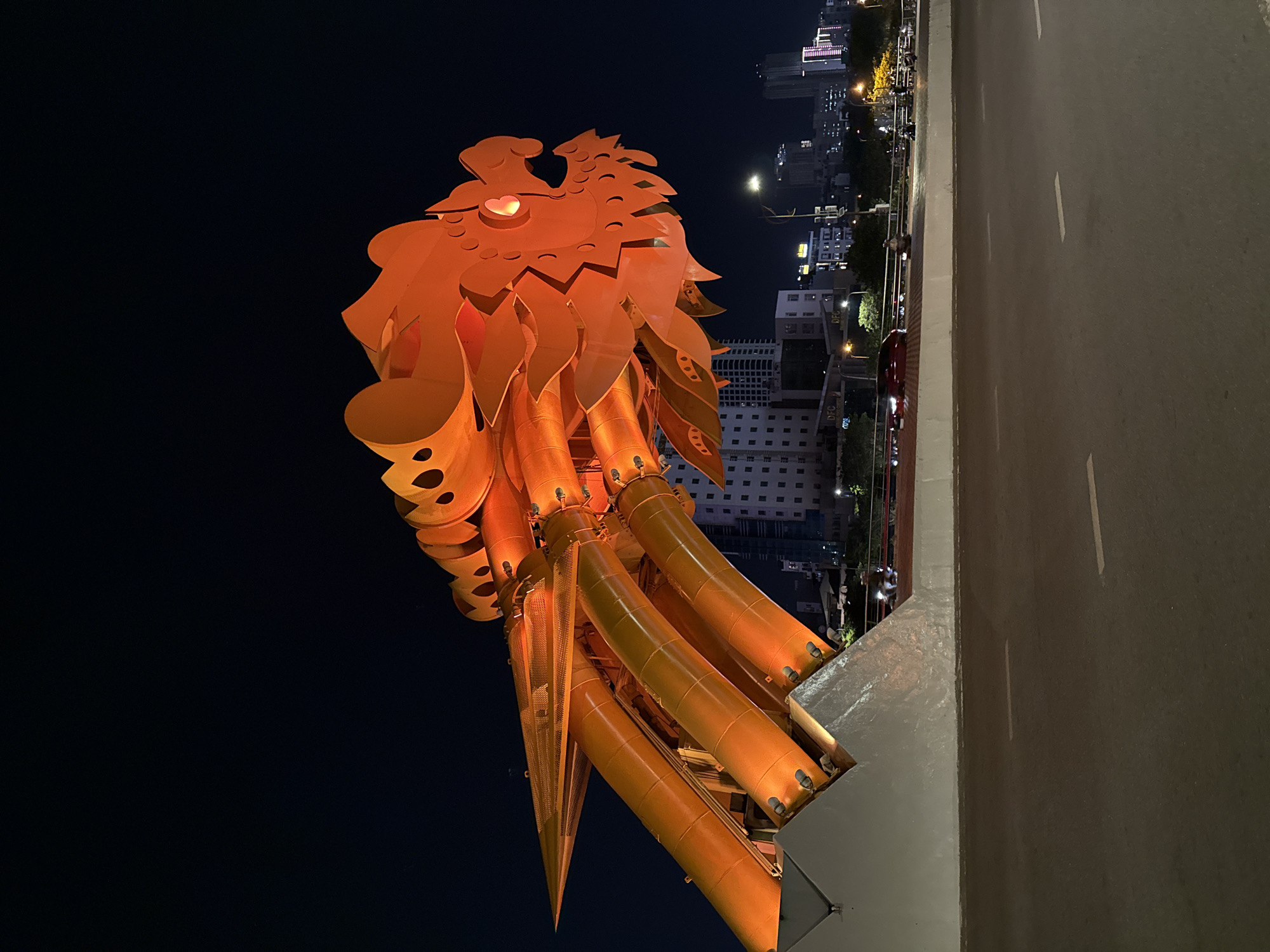 The image presents a striking nighttime view of the iconic Dragon Bridge in Đà Nẵng, Vietnam. The central subject is the highly stylized head and upper body of the dragon sculpture, illuminated in a vivid orange-gold color against a dark night sky. The dragon's head features intricate, angular elements resembling scales and a bright, glowing point that serves as an eye. Its neck and lower structure are composed of large, cylindrical tubes, adding to its monumental appearance.
In the background, to the right of the dragon, a dense urban landscape of Đà Nẵng is visible. Numerous multi-story buildings, many with illuminated windows, stretch into the distance, showcasing the city's modern architecture. Streetlights dot the scene, casting light on the surrounding area. Below the dragon, a robust section of the bridge structure, constructed from concrete and metal, is apparent. To the far right, a wide, paved road with distinct white lane markings indicates an active thoroughfare, likely part of or adjacent to the bridge.
The scene captures the Dragon Bridge as a prominent, illuminated landmark after sunset, emphasizing its artistic and structural grandeur within its urban setting. No people or specific events are depicted, and no visible text is discernible. The image is taken from a slightly elevated perspective, enhancing the dramatic presence of the dragon against the city lights.