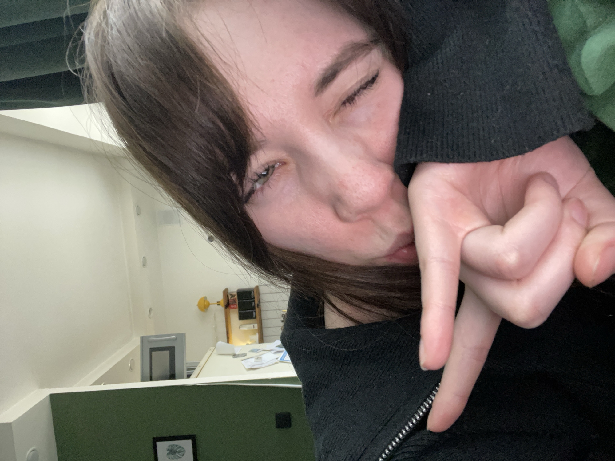 A selfie of a fair-skinned person with long, dark hair, one eye closed, and making a "love" hand gesture. The person is wearing a black zipped-up jacket. Behind the person is a room with white walls, a green section, and a desk. There are miscellaneous items on the desk, including what appears to be a small cabinet. The image does not contain sufficient information to confirm a location in Chaikovsky, Russia.