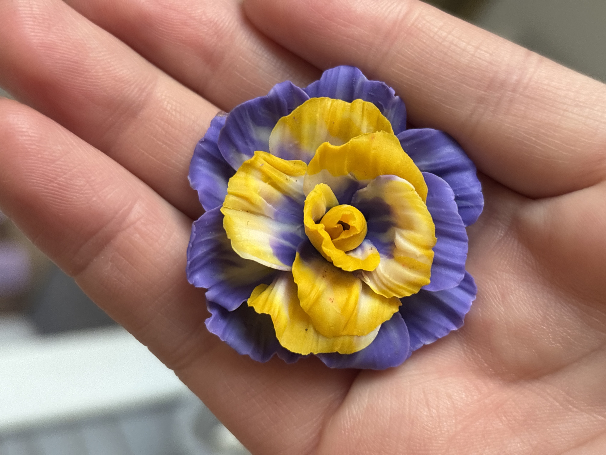 A close-up shot captures a decorative flower made of purple and yellow material resting in the palm of a hand. The flower is intricately designed with multiple layered petals. The hand, which appears to be that of an adult, is lightly cupped, showcasing the texture of the skin and the lines on the palm. The background is softly blurred, suggesting an indoor setting, with muted colors that do not distract from the main subject. No specific location in Blagoveshchensk, Russia, can be identified from the visual information provided.