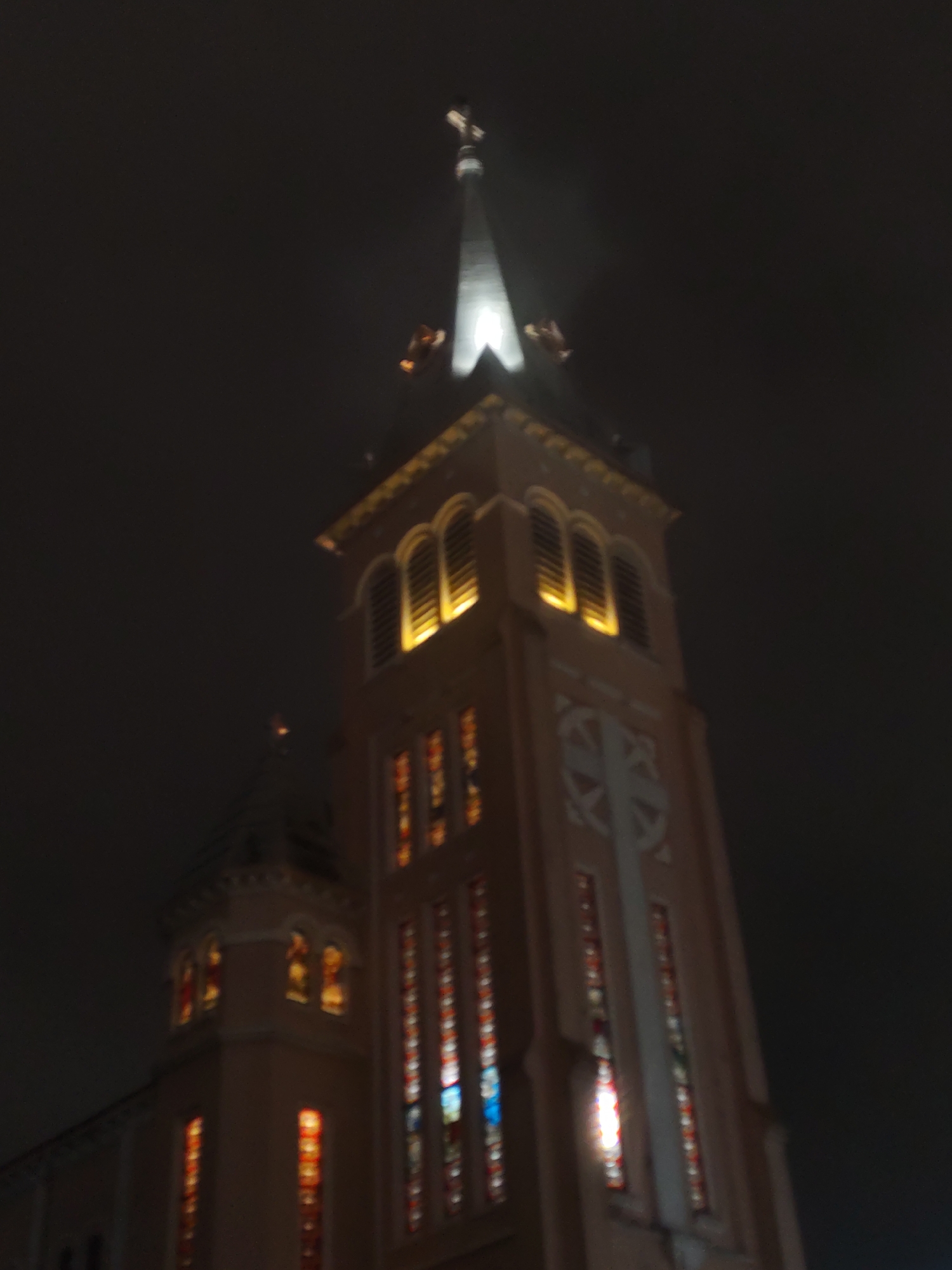 The image shows a night-time view of the Saigon Notre-Dame Basilica in Ho Chi Minh City, Vietnam. The basilica is a prominent landmark and a popular tourist attraction. The image is taken from a low angle, looking up at the basilica's spire. The spire is lit up, and the surrounding area is dark. There are no people or other objects visible in the image. The basilica is made of red brick and has a tall, pointed spire. The spire is topped with a cross. The image is slightly blurry, possibly due to low light conditions or movement of the camera.