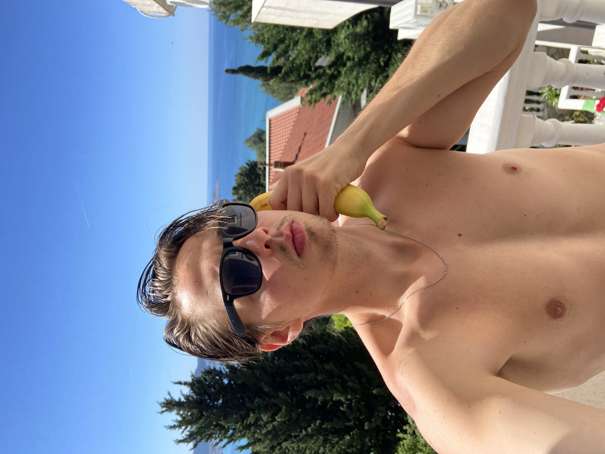 A shirtless man with dark sunglasses is holding a banana to his ear, posing as if it's a telephone, with a sea and blue sky in the background. He has wet hair, a silver chain around his neck, and light skin. The location appears to be Šušanj, Montenegro, with a view of the Adriatic Sea, trees, and a red-roofed building. A white balcony railing is also visible in the background.