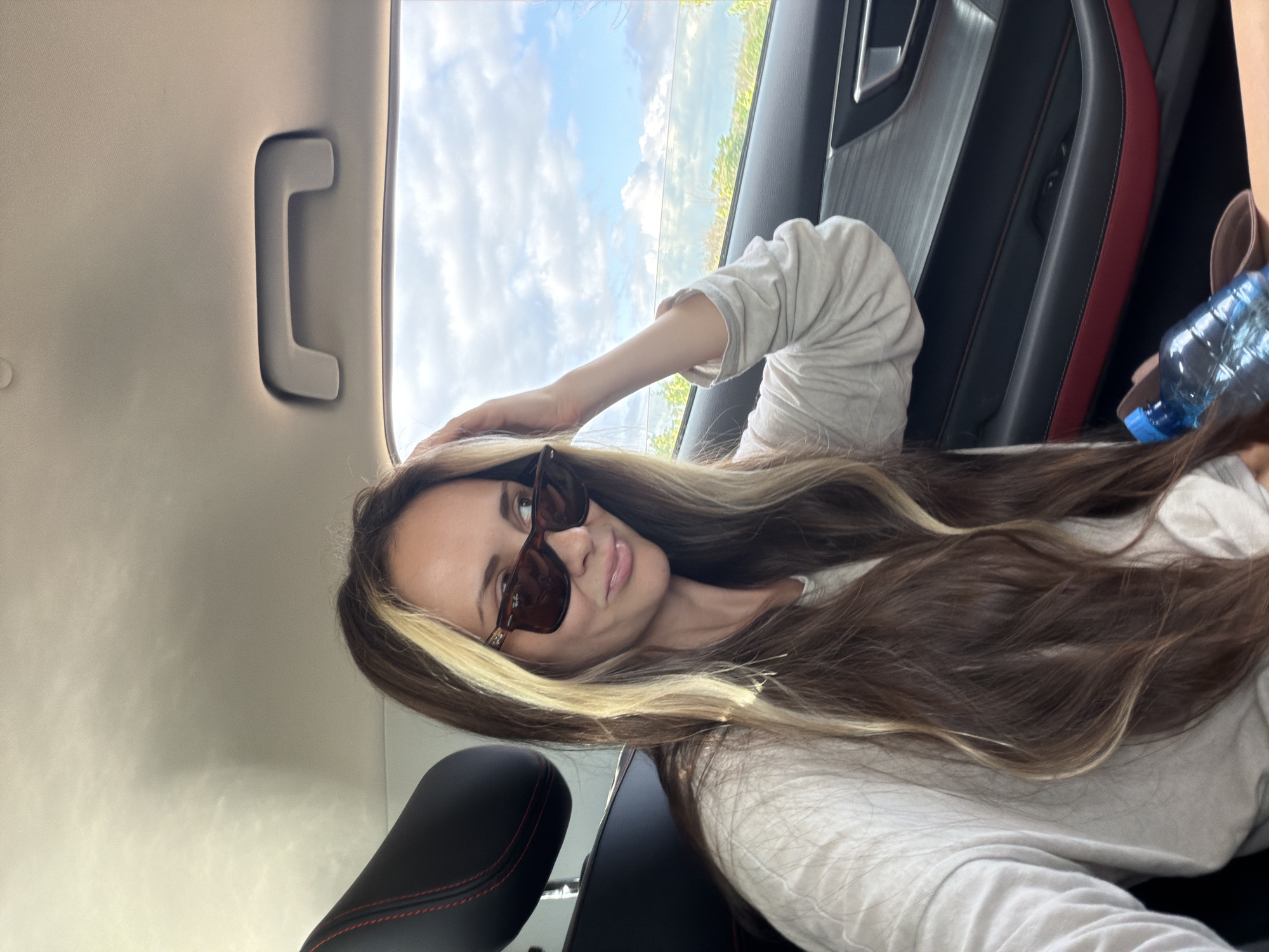 A woman with long brown hair with blonde highlights, wearing sunglasses and a light-colored long-sleeved shirt, is sitting in the back seat of a car. She is touching her hair with her right hand and looking to her right. A blue water bottle is visible in the lower right corner of the image. The interior of the car features black seats with red stitching and a metallic gray trim on the door panel. Outside the car window, a partly cloudy sky and green landscape are visible. The image was taken in Ternovka, Russia.