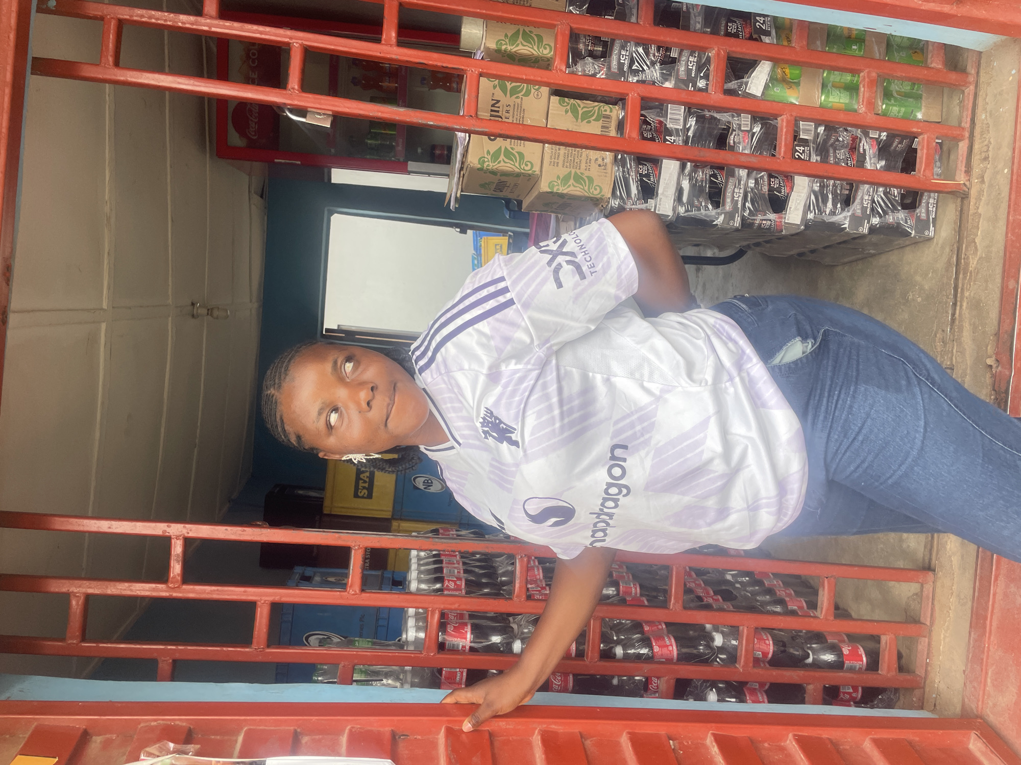 A young woman stands in front of a small shop in Jos, Nigeria. The shop's metal security gate, painted red, is pulled down and open. Behind the gate, shelves are visible stocked with drinks like Coca-Cola, Guinness, and other bottled beverages, as well as boxes of goods. The woman wears a white and lavender Manchester United jersey with "Snapdragon" printed on the front, and blue jeans.