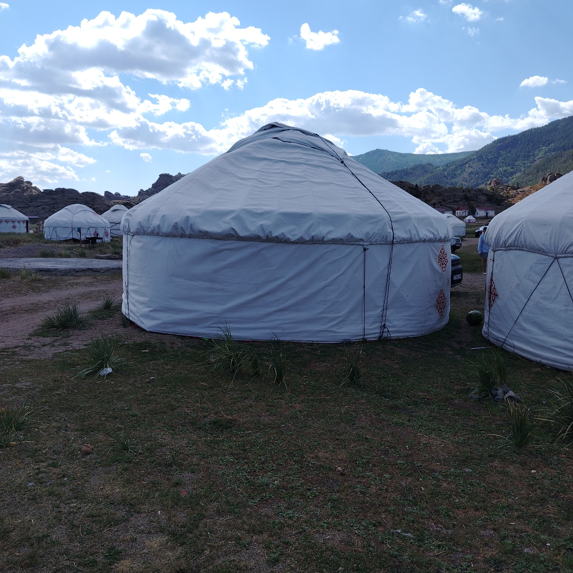 The image depicts a collection of yurts, traditional Mongolian dwellings, situated in an outdoor environment. The yurts are predominantly white with some displaying red decorative patterns on their exterior. They are arranged in a cluster, with some in the foreground and others further in the background. The ground around the yurts is a mix of dry grass and dirt. In the background, rolling hills covered with greenery and rocky outcrops are visible, under a partly cloudy sky with a clear blue sky dominating. A few vehicles, including a car, are parked near the yurts. There are also a few people visible, one of whom is standing between two yurts in the middle ground, facing away from the camera. The overall scene suggests a rural or nomadic settlement, possibly a tourist campsite, in the region of Торайғыров, Kazakhstan.