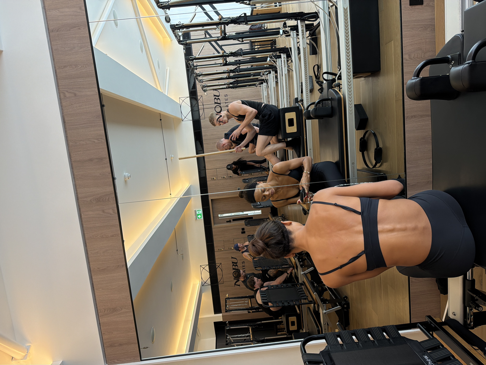 The image shows a group of people in a brightly lit Pilates studio, as seen through a large mirror. In the foreground, a woman with her back to the camera is seated on a Pilates reformer, wearing a black sports bra and leggings. Her hair is styled in a bun. The mirror reflects the rest of the studio and its occupants.
In the reflection, several individuals are engaged in Pilates exercises. Two men are seated on reformers, one of whom is holding a wooden stick and looking at his phone. Another man is actively adjusting equipment on a reformer. A woman is also seen in the reflection, sitting on a reformer with her legs extended. The studio features multiple Pilates reformers arranged in rows, with exercise equipment like straps and handles visible. The walls are paneled with wood, and the ceiling has recessed lighting. The "NOBU PILATES" logo is prominently displayed on the back wall. The overall environment appears clean and modern, suggesting a high-end fitness facility. The presence of the "NOBU PILATES" branding indicates the location is likely a Nobu Pilates studio, which has locations globally, including in London, United Kingdom.