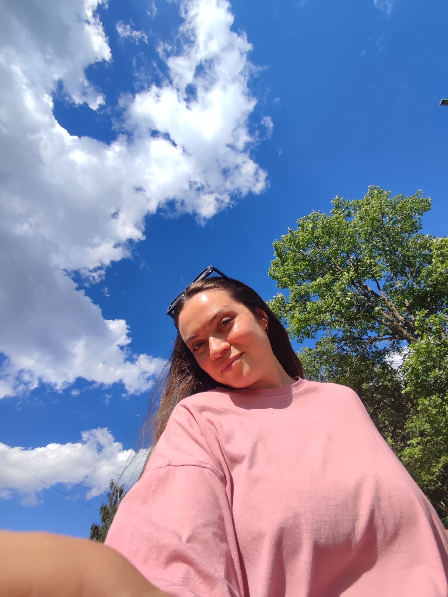 This media file is a selfie taken outdoors on a sunny day with a bright blue sky and scattered white clouds. A young woman with long brown hair is the primary subject, wearing a pink t-shirt and sunglasses perched on her head. She is smiling gently at the camera, suggesting a relaxed and happy mood.
Behind her, a large, leafy green tree dominates the upper right portion of the frame, indicating a park or natural setting. The sunlight creates a warm glow on her face and highlights the fabric of her shirt. The low-angle perspective emphasizes the vastness of the sky above. The overall scene conveys a sense of summer leisure and enjoyment. No text is visible in the image. The location context is Chaikovsky, Russia.