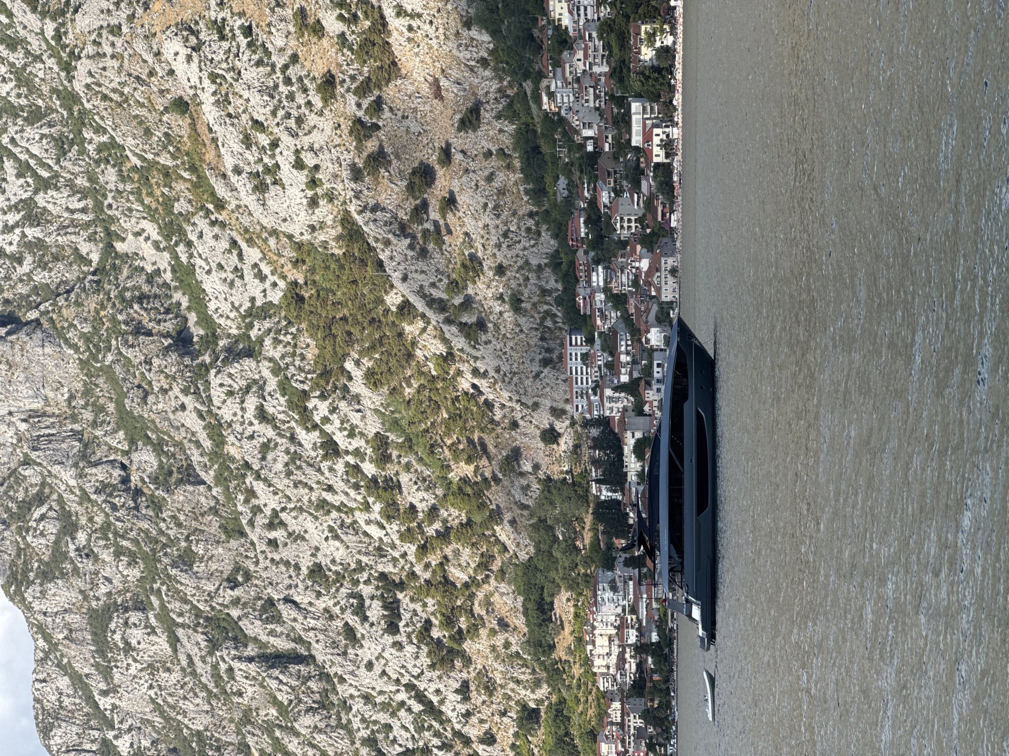 A superyacht with a tender boat alongside is moored in the Bay of Kotor, near the coastal town of Škaljari, Montenegro. The yacht is dark gray and sleek, with a streamlined hull and a multi-level superstructure. The tender is smaller and white, positioned to the port side of the yacht. In the background, a dense cluster of residential buildings with red-tiled roofs climbs a steep, rocky mountainside dotted with green vegetation. The water in the bay is calm and murky. The sky is partly cloudy.