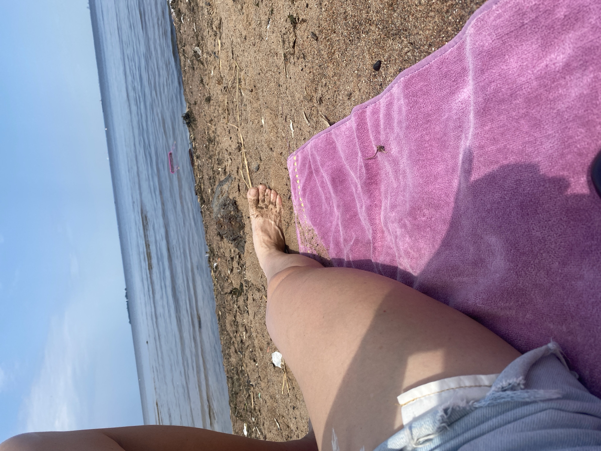 A person's legs are visible, resting on a pink towel on a sandy beach. The person is wearing denim shorts. The water of the Gulf of Finland stretches out behind them, with a hazy horizon and a few distant ships. There is some debris on the beach, including sticks and what appears to be a pink plastic object floating in the water. The scene suggests a sunny day at the beach in Saint Petersburg, Russia.