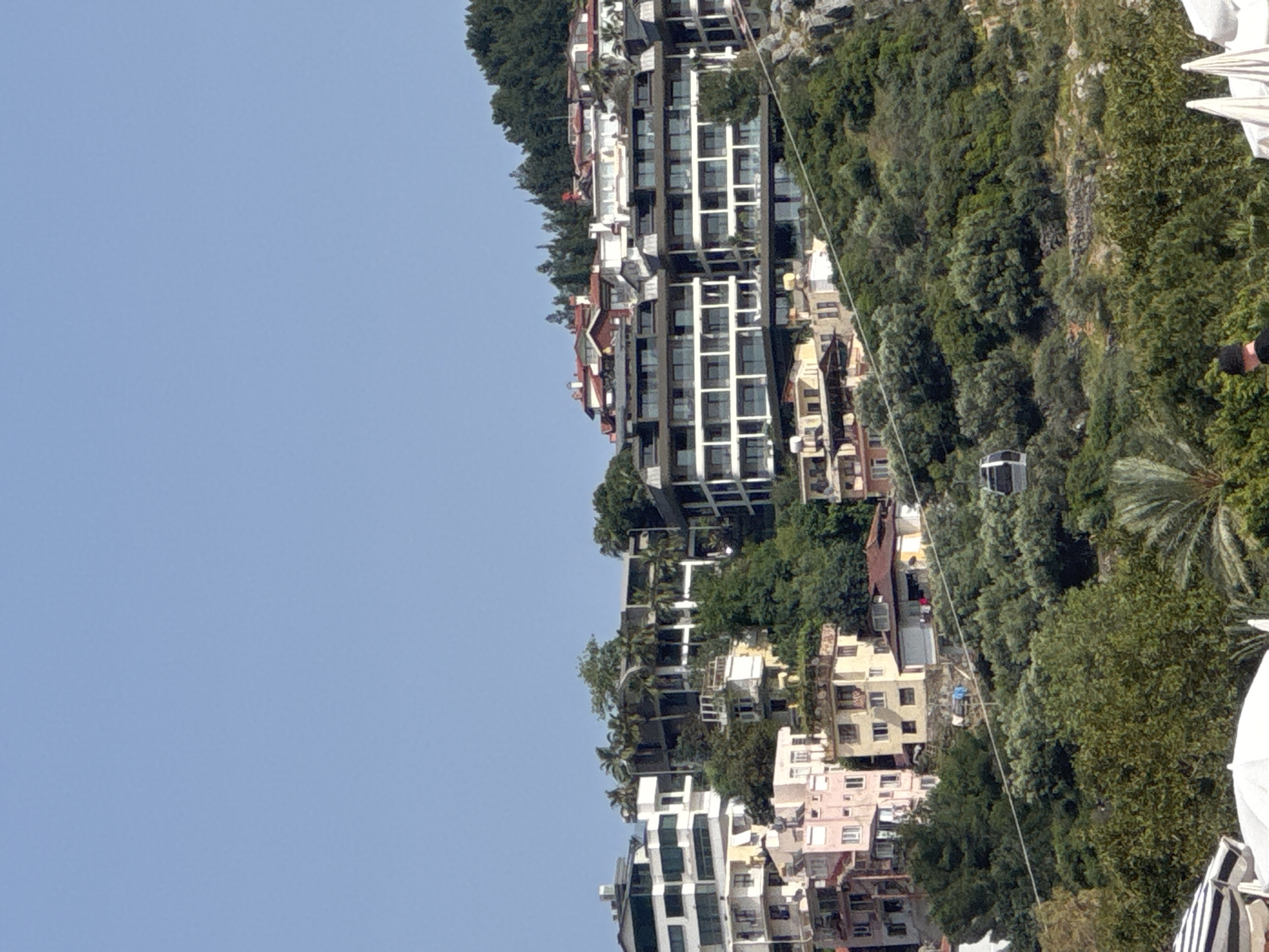 A hillside view shows a collection of multi-story buildings, including modern apartment complexes with balconies, interspersed with trees. A cable car system is visible, with a cabin positioned between trees. The sky is clear and blue. This is likely a view of the urban landscape on a hillside in Alanya, Turkey.