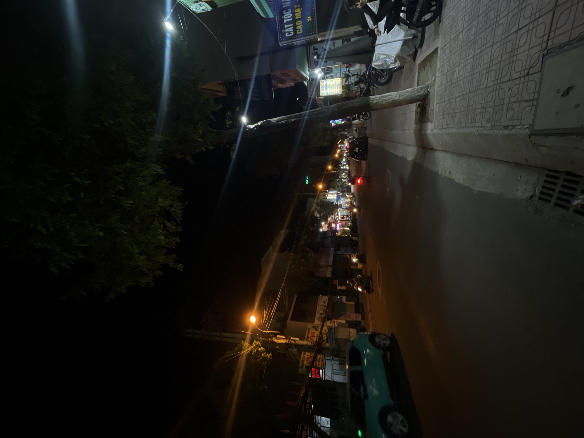The image depicts a night scene on a street in Bac Nha Trang Ward, Vietnam. The street is lined with businesses and illuminated by streetlights and the lights from the establishments. A turquoise car is visible on the left side of the road, and several motorcycles are on the road and parked on the sidewalk. Trees with green foliage are present along the street. The sidewalks are paved with patterned tiles. The overall impression is of a busy urban environment at night.