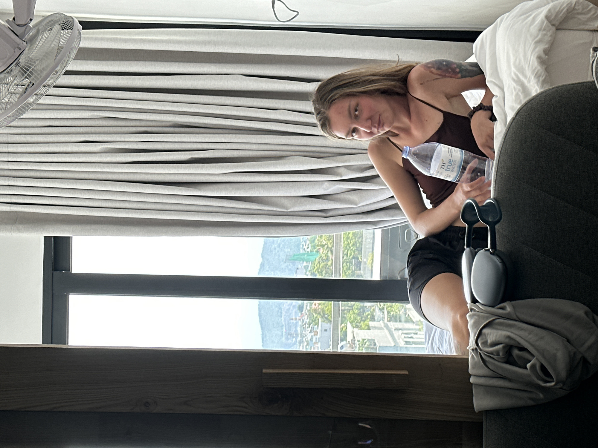 A young woman with long brown hair and a tattoo on her left arm is lying on a bed in a room with a view of the city of Đà Nẵng, Vietnam. She is wearing a dark brown tank top and black shorts. She is holding a bottle of water. To her left, on the bed, are a pair of headphones and a folded gray shirt. In the background, through a large window, a cityscape with buildings and mountains can be seen. A ceiling fan is visible in the upper right corner of the image. The room appears to be a hotel room.