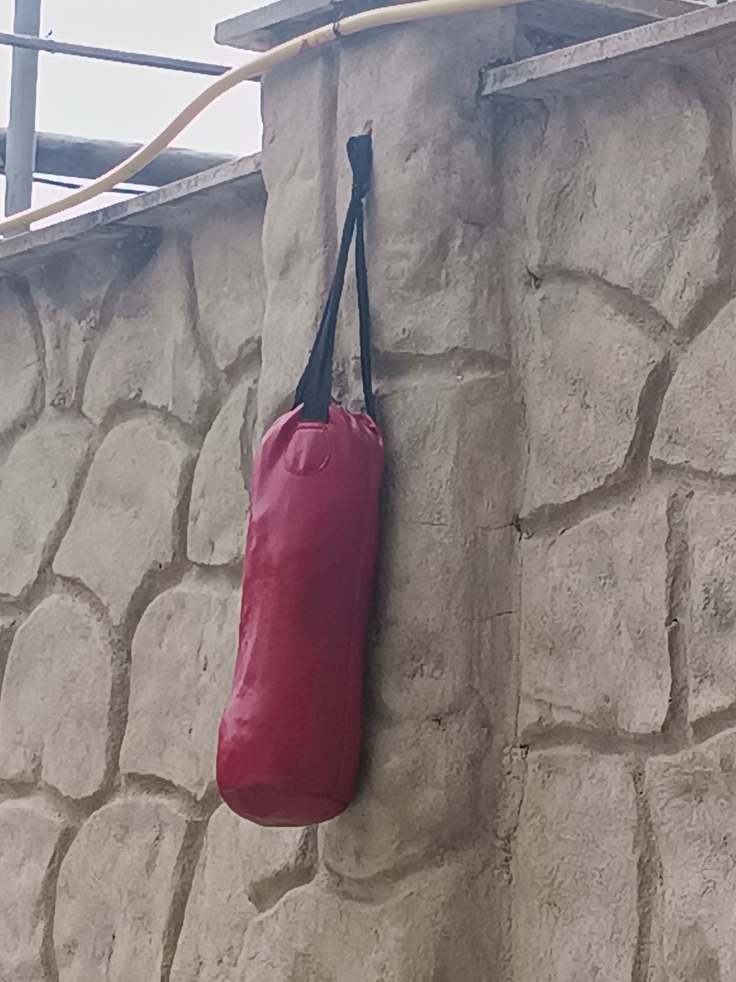 A red punching bag is hanging from a hook on a stone pillar. The pillar is part of a wall made of rough, irregularly shaped stones. The wall appears to be outdoors, with a cloudy sky visible in the background. A yellow pipe runs horizontally above the wall. The scene is likely in Tbilisi, Georgia.