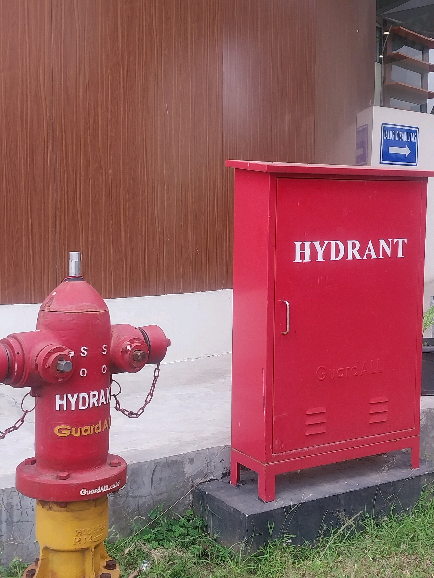 In Sleman, Indonesia, a red fire hydrant with a yellow base stands on a concrete pad next to a red metal cabinet. The hydrant has several outlets and chains attached. The cabinet is labeled "HYDRANT" in white letters and "GuardAll" embossed at the bottom. A blue sign with white text and an arrow, indicating "JALUR DISABILITAS" (disability lane), is visible in the background on a white structure. The scene is situated against a wooden-paneled wall and a grassy area.