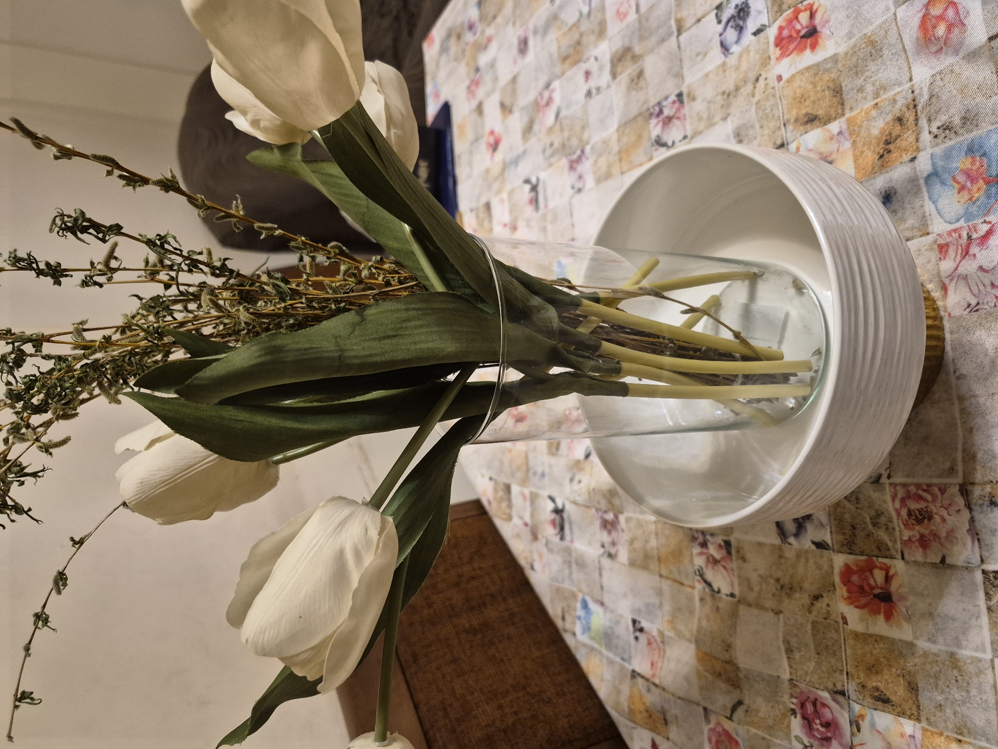 This close-up, rotated image, taken in Yerevan, Armenia, displays an indoor decorative floral arrangement. The main subjects are several artificial white tulips with prominent green leaves and stems. They are accompanied by dried green foliage, which includes slender branches with small buds or catkins, and some wiry, darker green elements.
These elements are contained within a clear, cylindrical glass vase. The vase is placed inside a larger, decorative white bowl, which has a ribbed exterior texture and an inward-curving rim. The entire arrangement rests on a patterned tablecloth. The tablecloth features a mosaic-like design composed of square and rectangular patches in various shades of beige, brown, and off-white, interspersed with vibrant floral motifs in pink, red, blue, and purple hues.
The image captures a still life, with no activity or event taking place. The lighting is soft and consistent, typical of an indoor environment, making the time of day indeterminate. No specific location cues beyond the given context are visible, and no emotions are conveyed by the inanimate subjects. There is no visible text within the image.